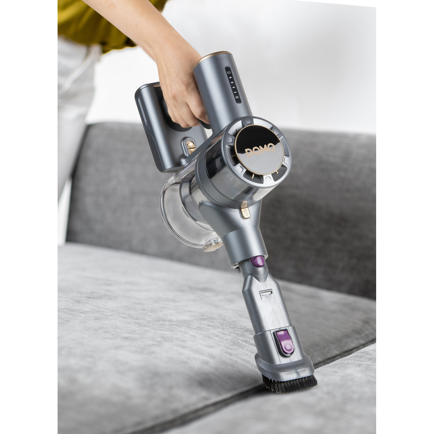 Personne utilisant un aspirateur gris sur un canapé gris. L'aspirateur a un bouton violet.