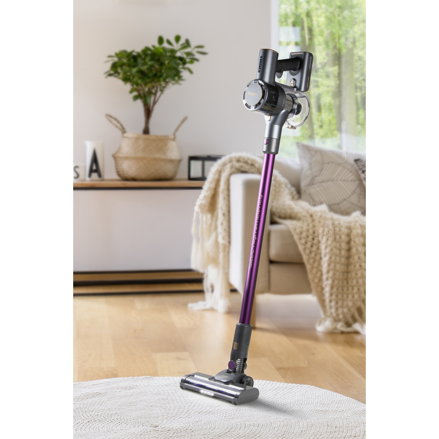 Un aspirateur sans fil violet et argenté est posé sur un tapis, avec une fenêtre et un canapé en arrière-plan.