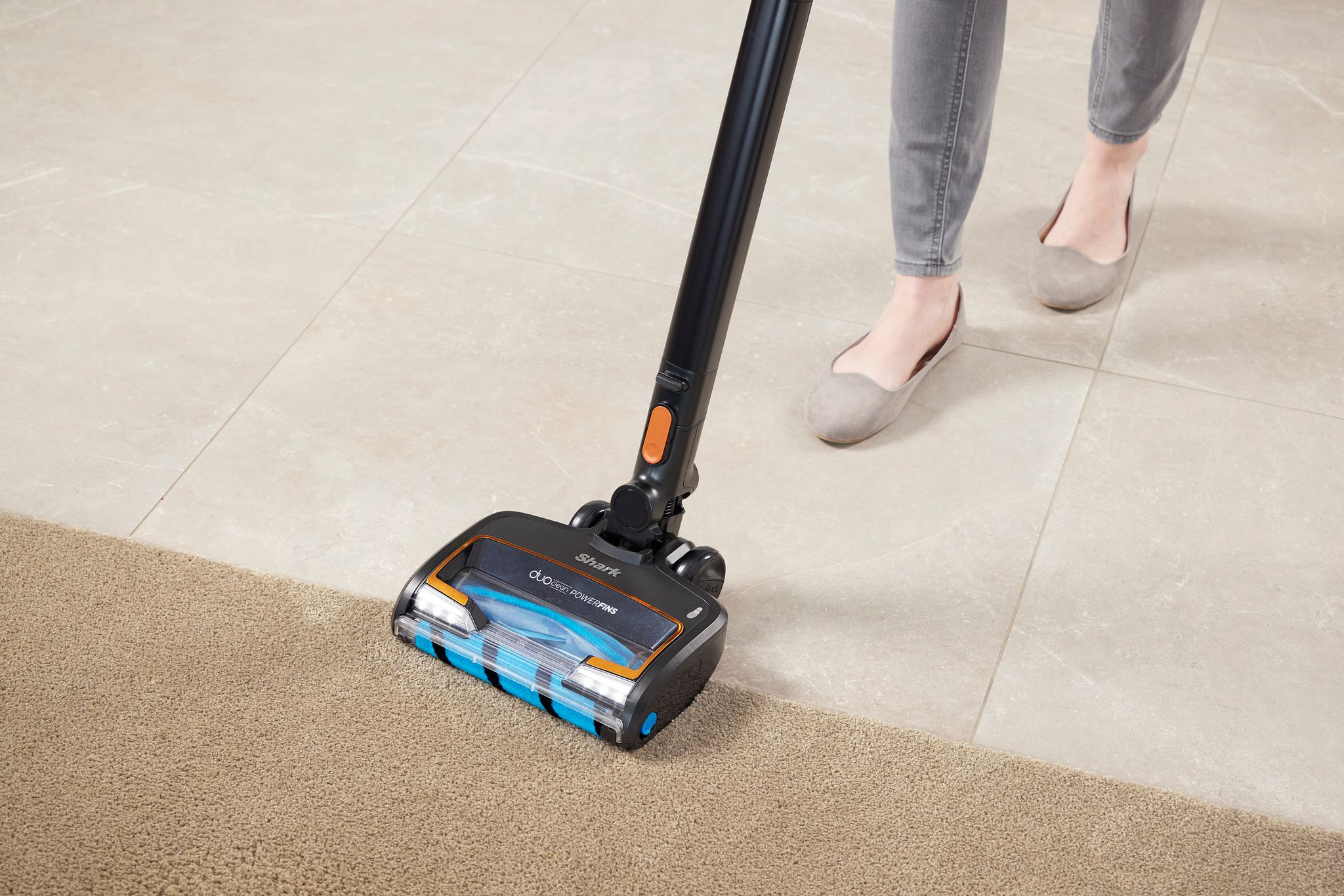Une personne passant l'aspirateur sur un tapis beige avec un aspirateur noir et orange sur un sol carrelé.