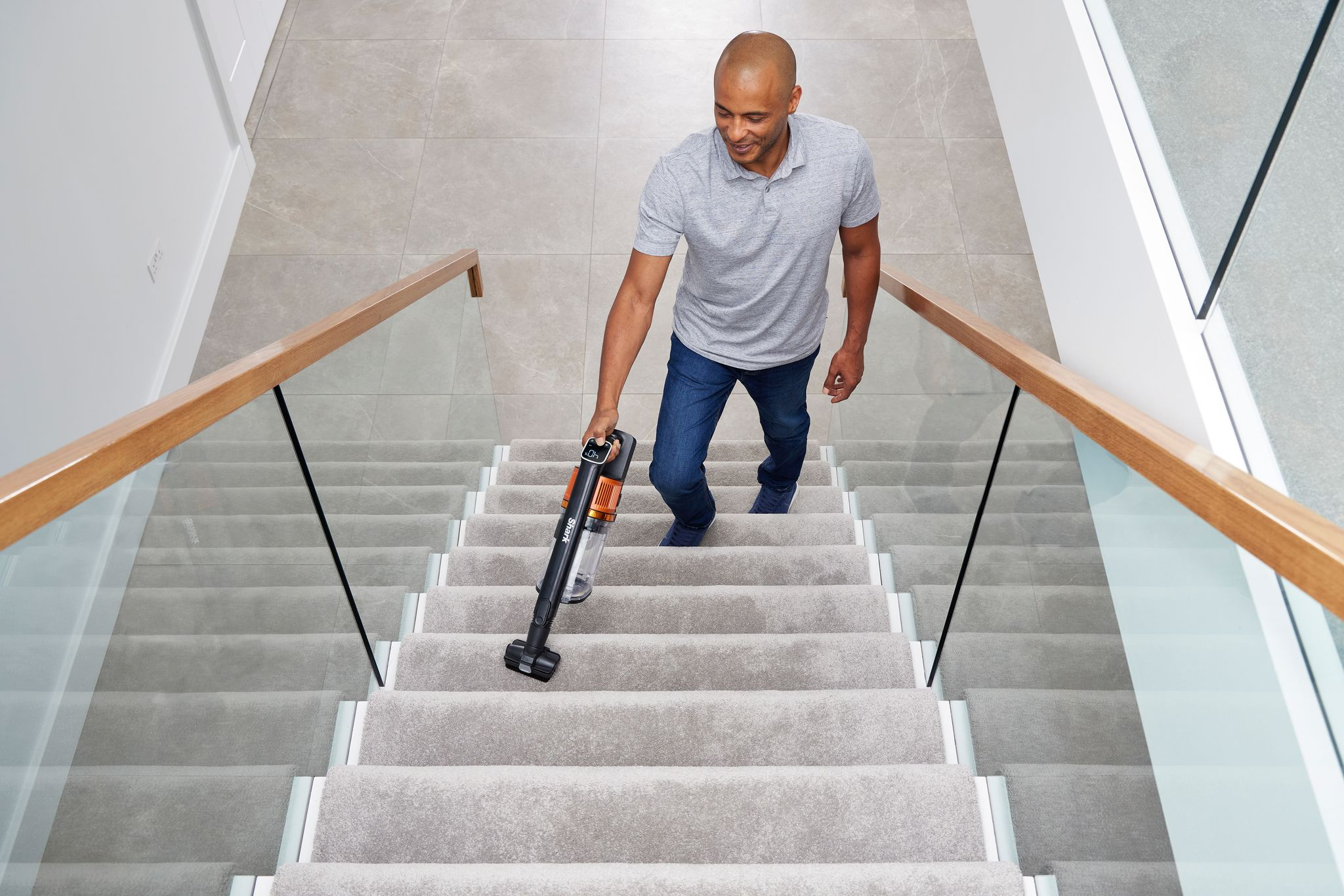 Homme passant l'aspirateur sur un escalier moquetté avec un aspirateur à main. Escalier avec rampes en verre.