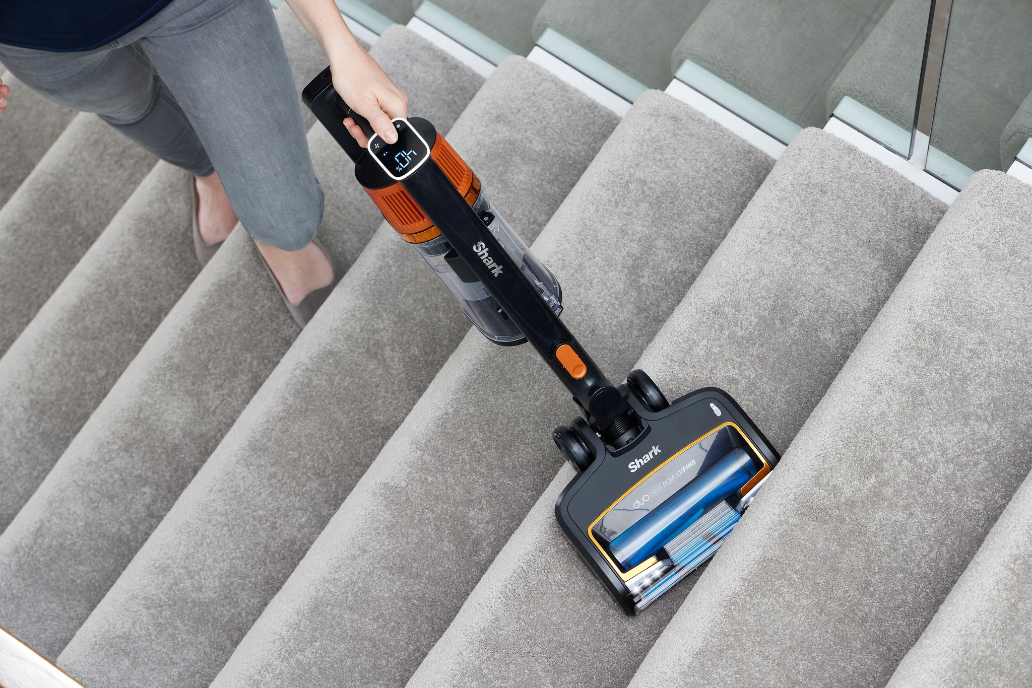 Une personne passant l'aspirateur sur un escalier recouvert de moquette avec un aspirateur Shark noir et orange.