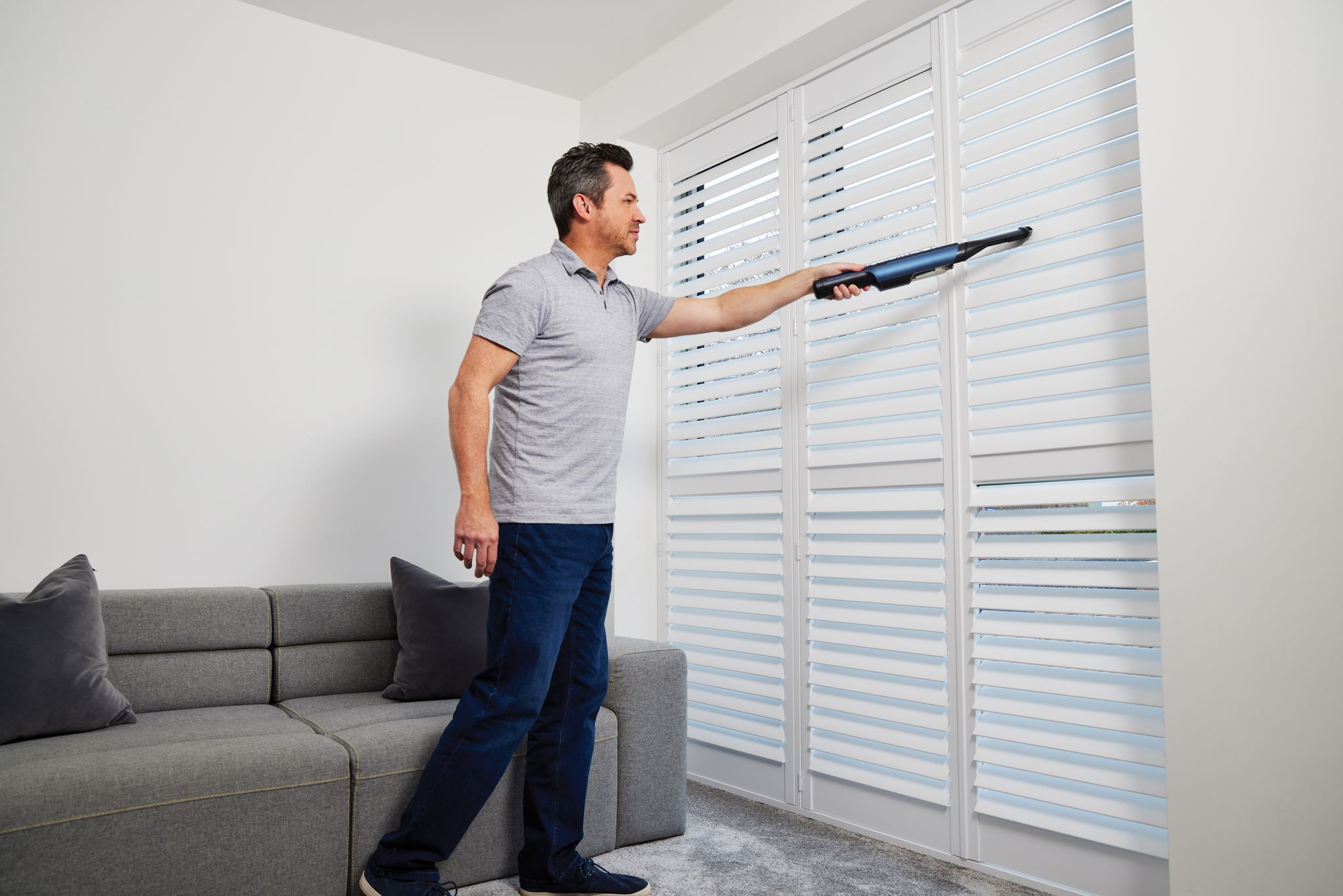 Ein Mann saugt weiße Fensterläden ab. Er trägt ein graues Hemd und Jeans.