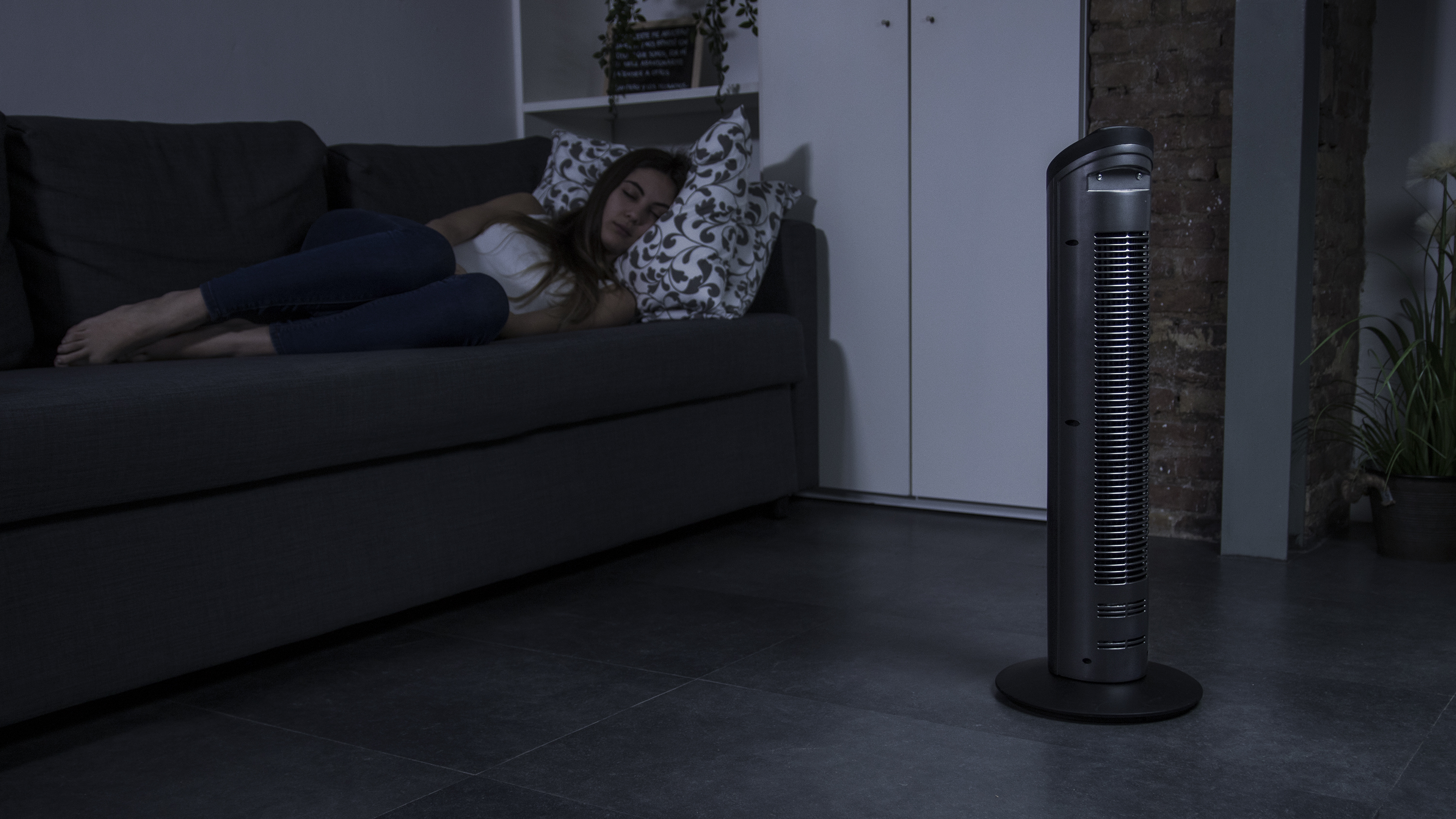 Una mujer duerme en un sofá gris. Un ventilador está junto al sofá en una habitación con poca luz.