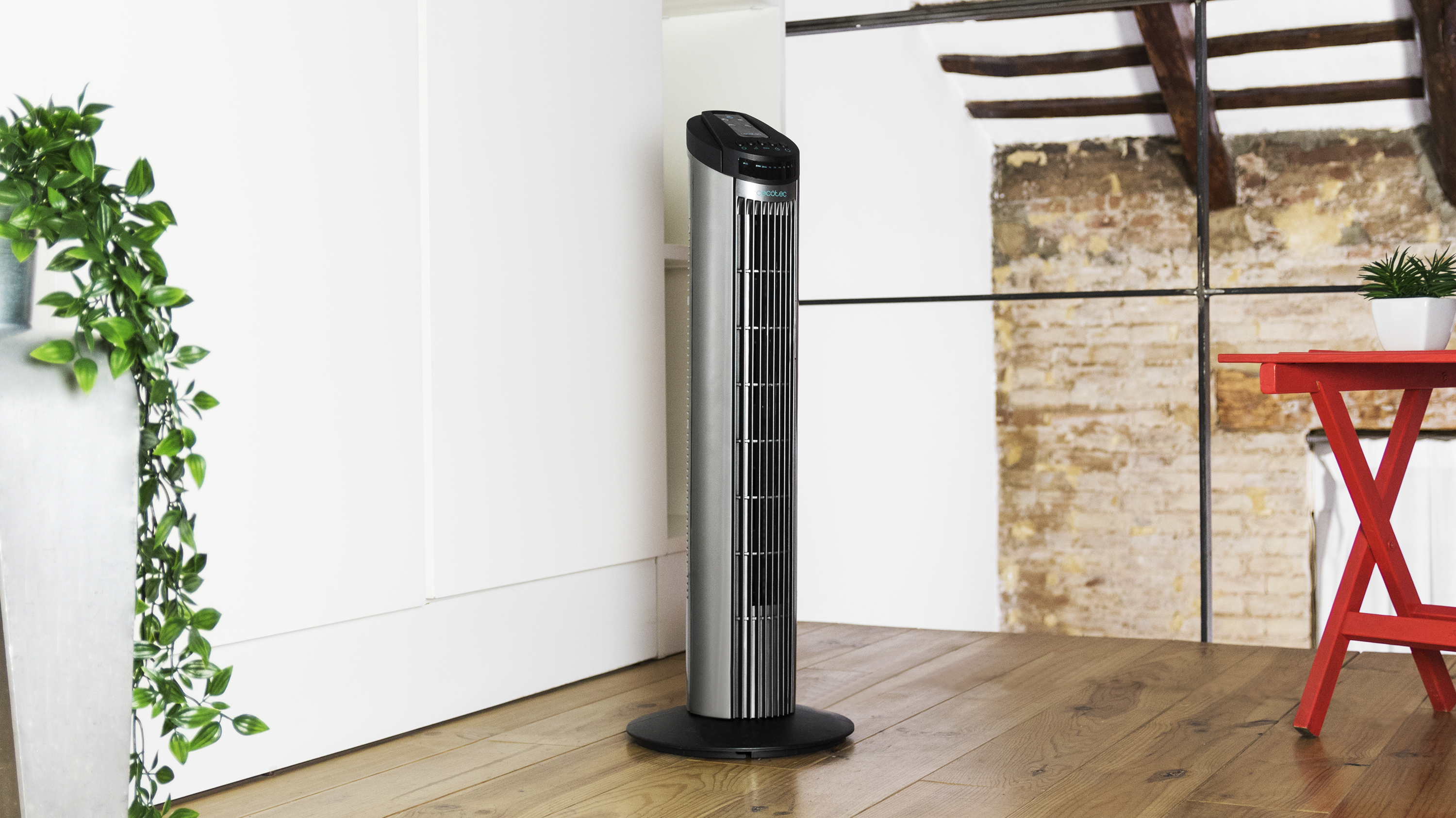 Ventilador alto plateado en una habitación. Está en un piso de madera. Plantas y mesa roja al fondo.