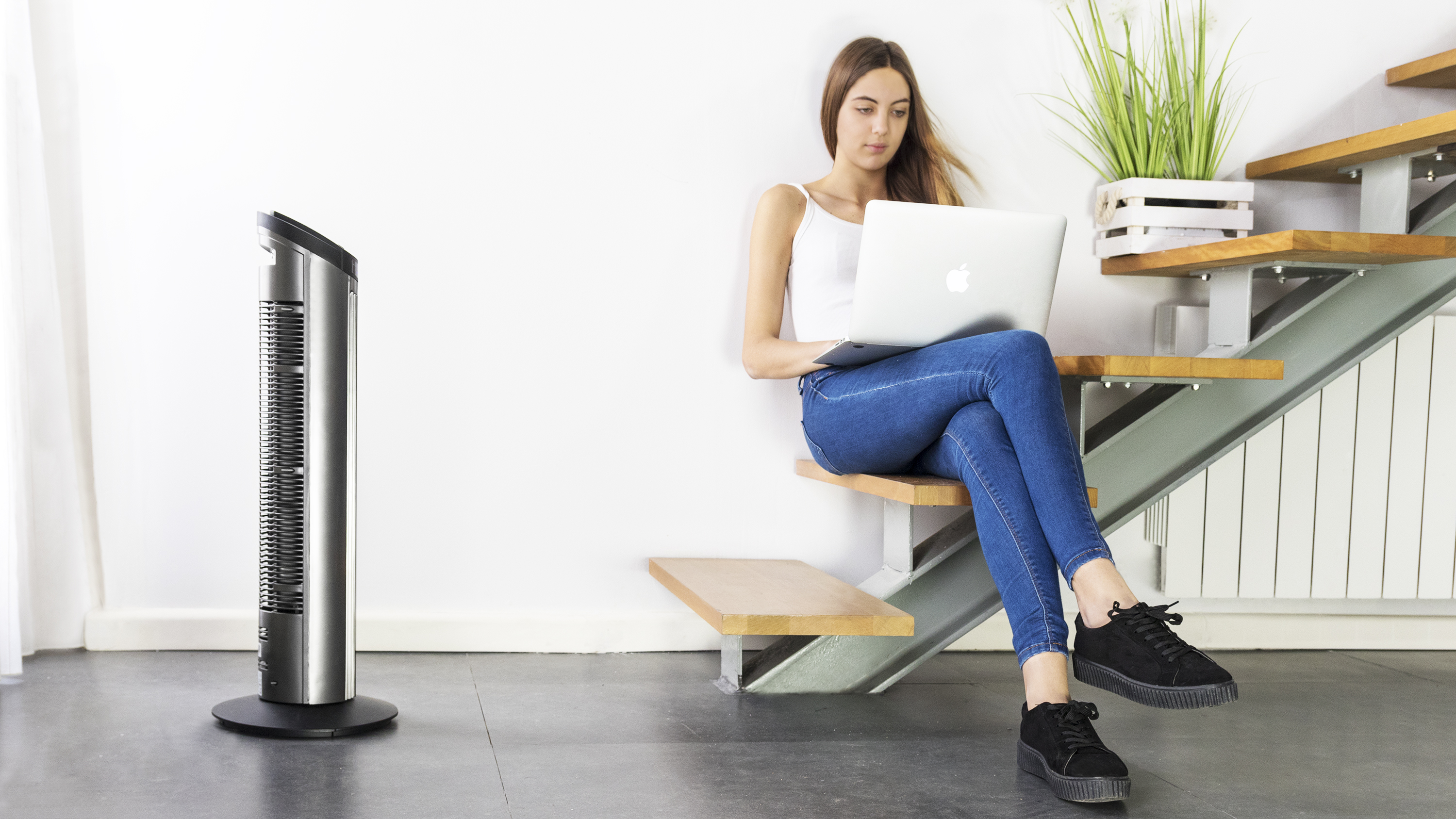 Mujer en escaleras con portátil y ventilador. Ventilador a la izquierda, escaleras y pared blanca.