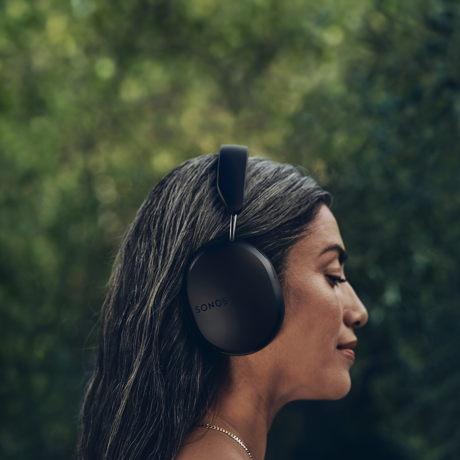 A woman with gray hair wearing black SONOS headphones against a blurred green background.