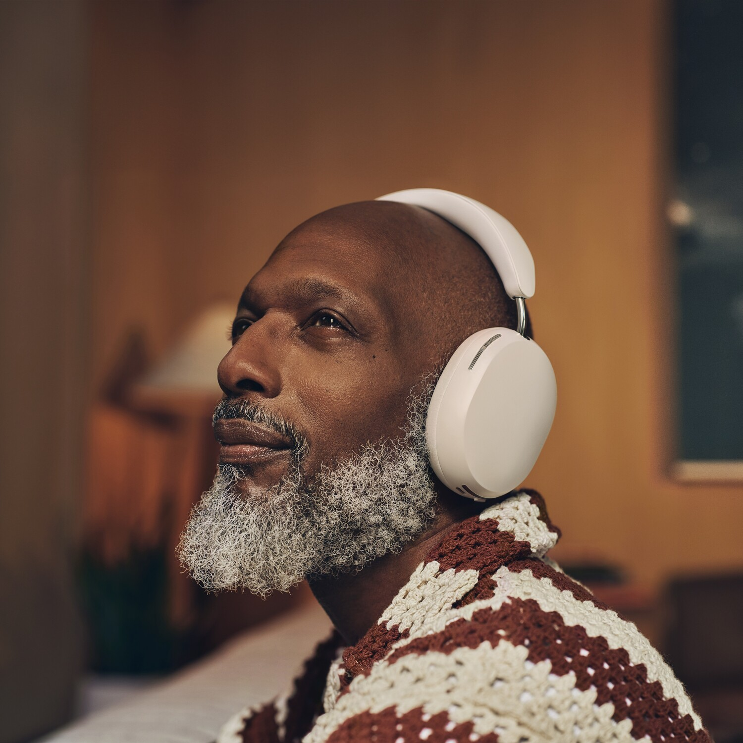 A man with a gray beard wearing white headphones looks up, sitting in a room with a brown wall.