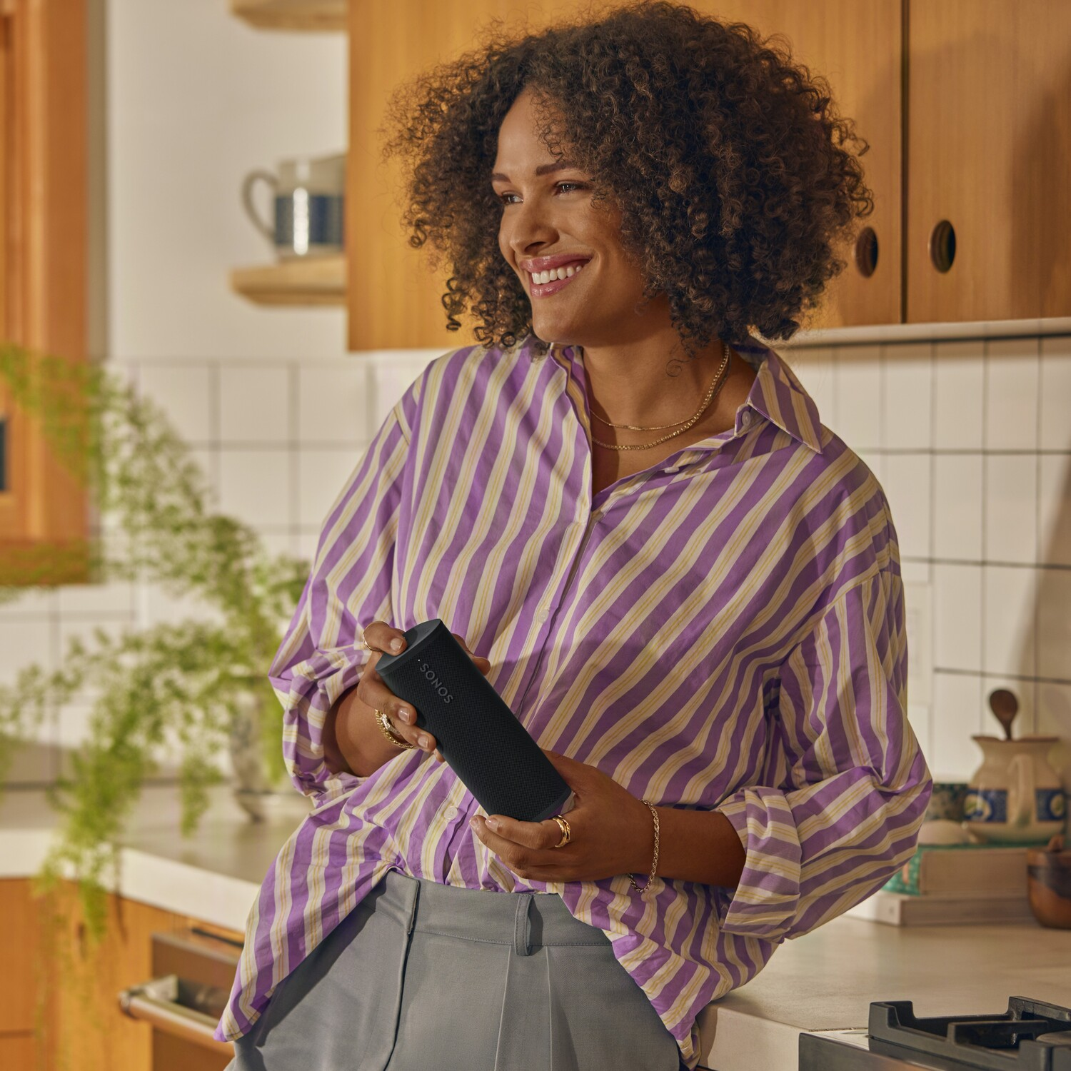 Vrouw houdt een zwarte luidspreker vast bij een keuken. Ze lacht en draagt een paars gestreept overhemd.
