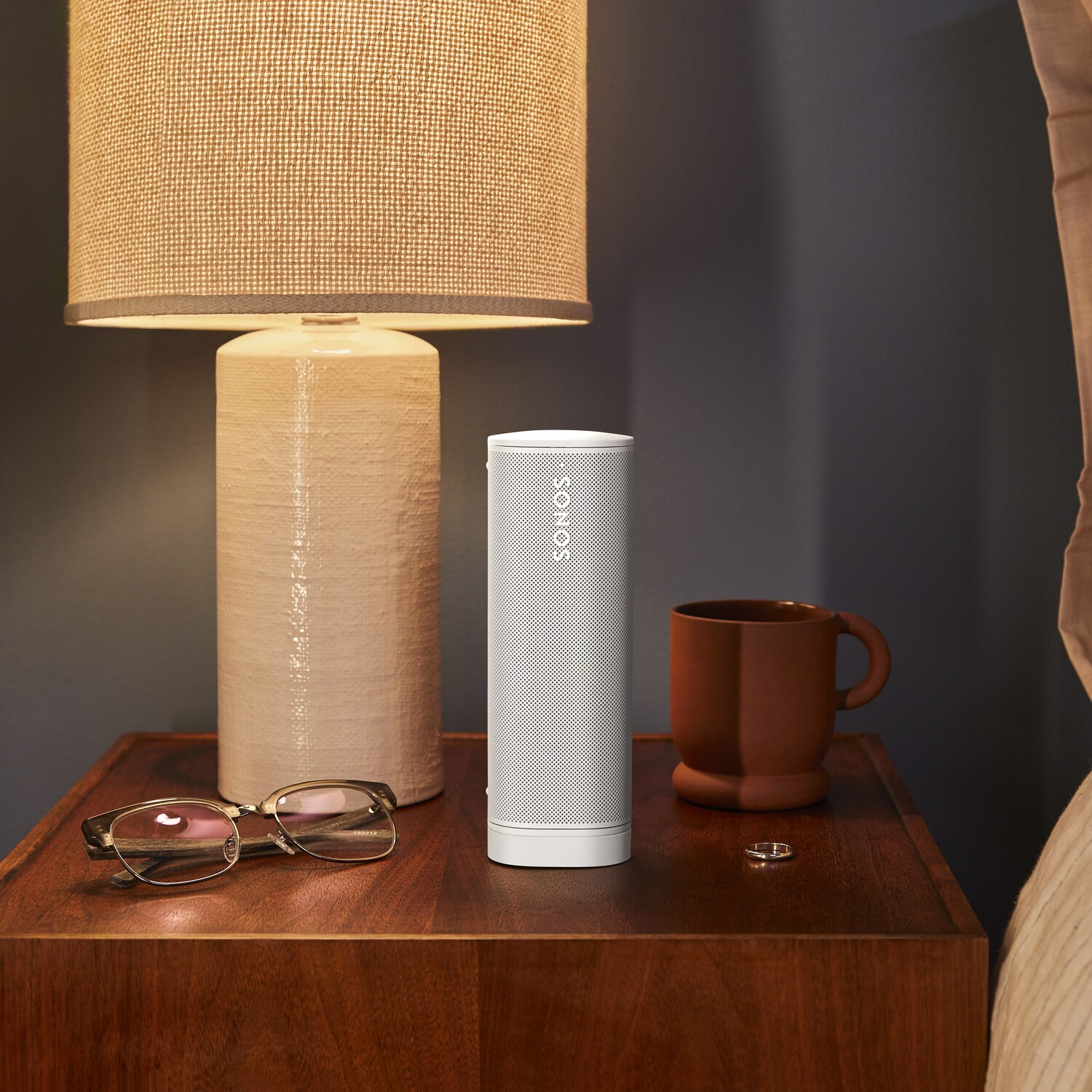 White speaker on a nightstand next to a lamp, glasses, and a coffee mug. The background is a bed.
