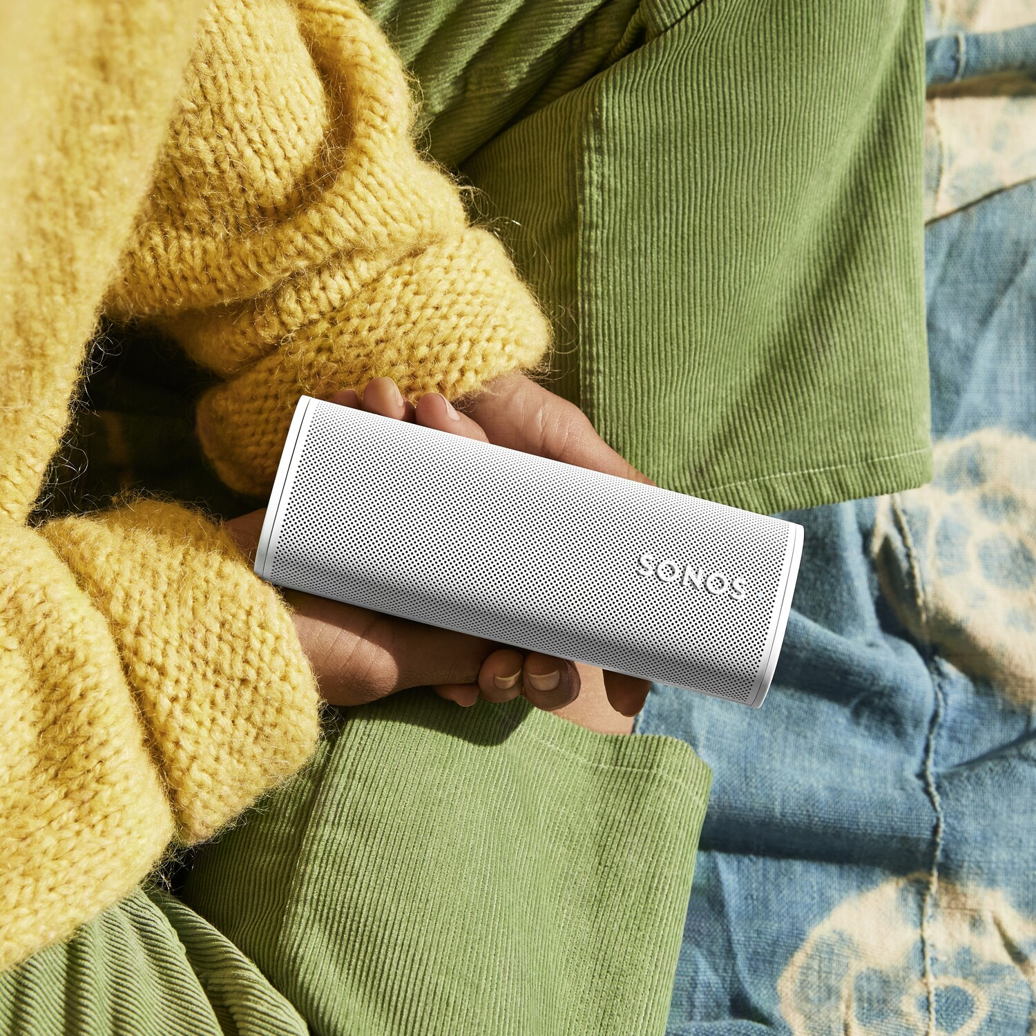A person holds a white Sonos speaker. They are wearing a yellow sweater and green corduroy pants.