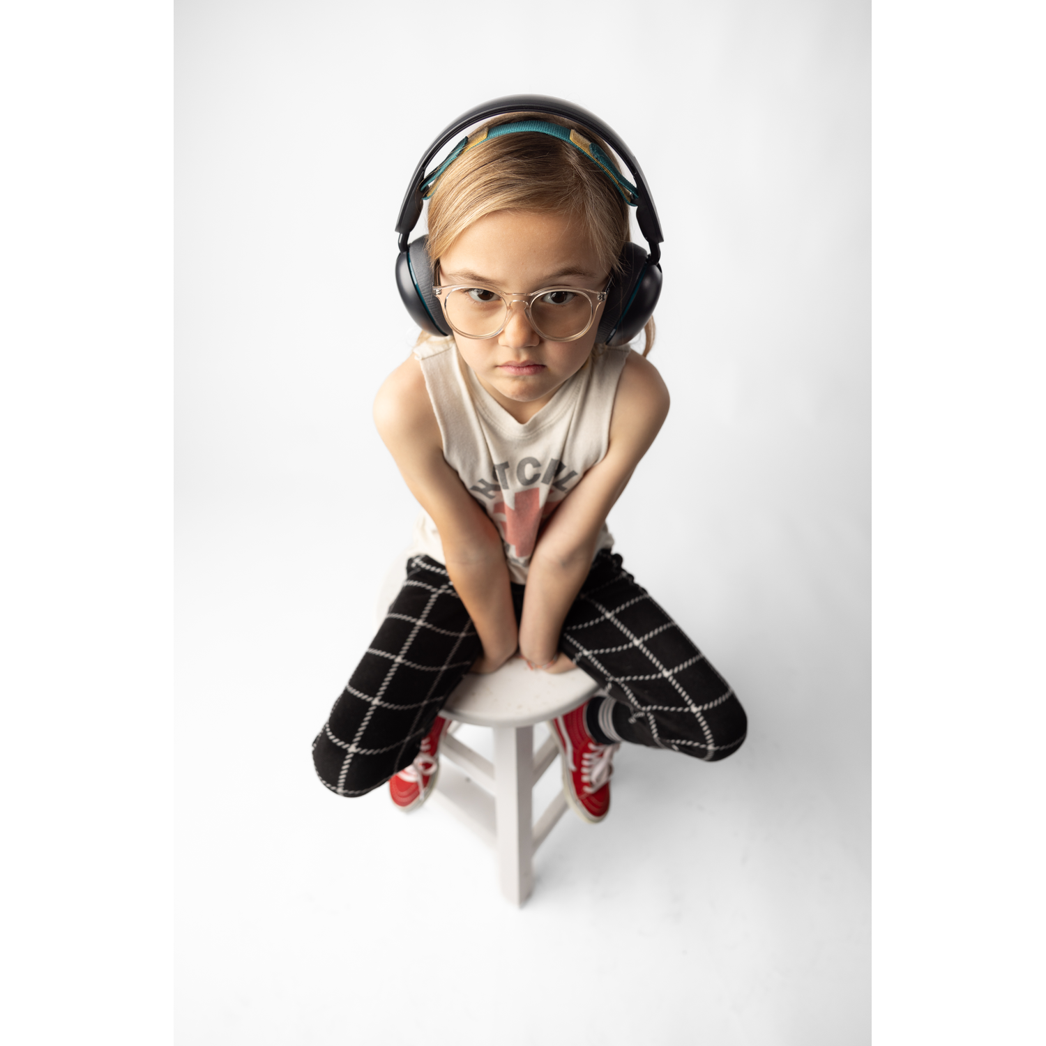 A young girl wearing glasses and headphones sits on a stool against a white backdrop, looking at the camera.