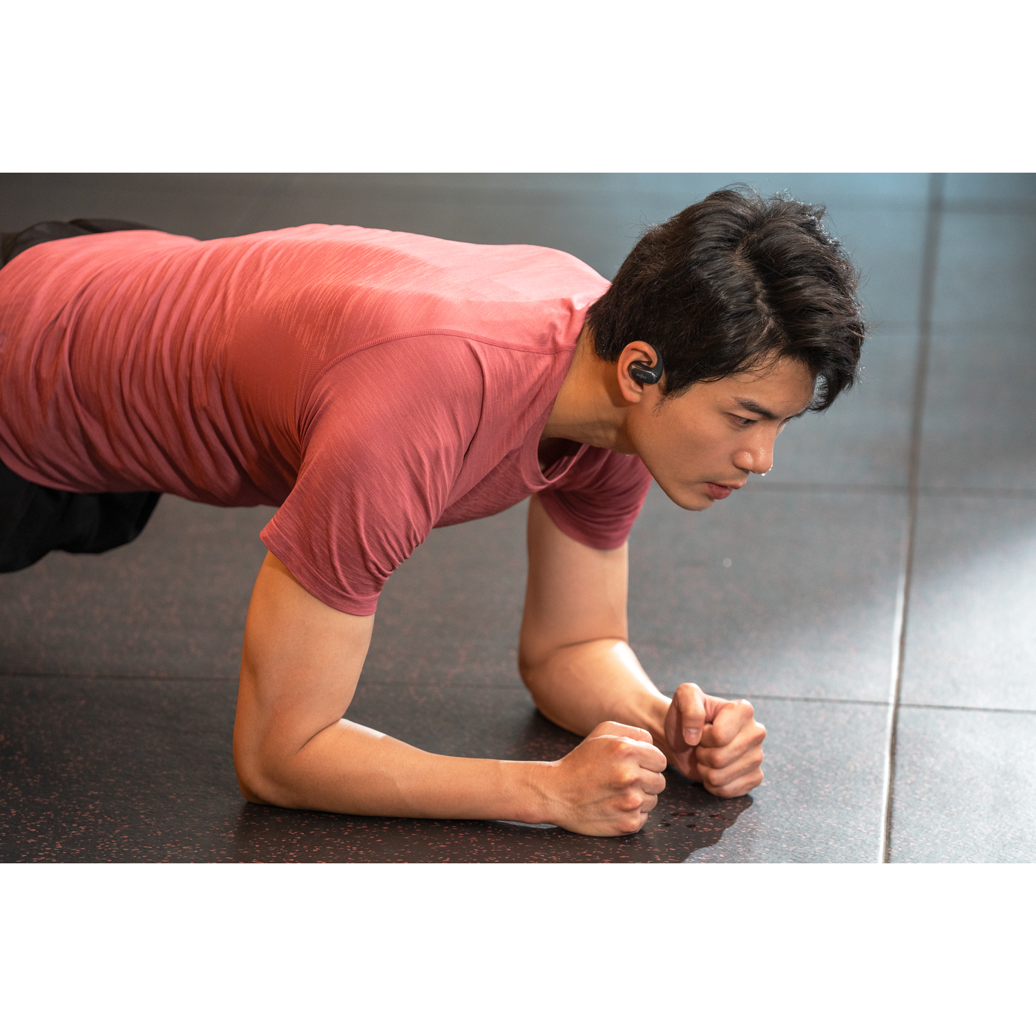 Man in plank position wearing earbuds, exercising on a gym floor. Focused expression.