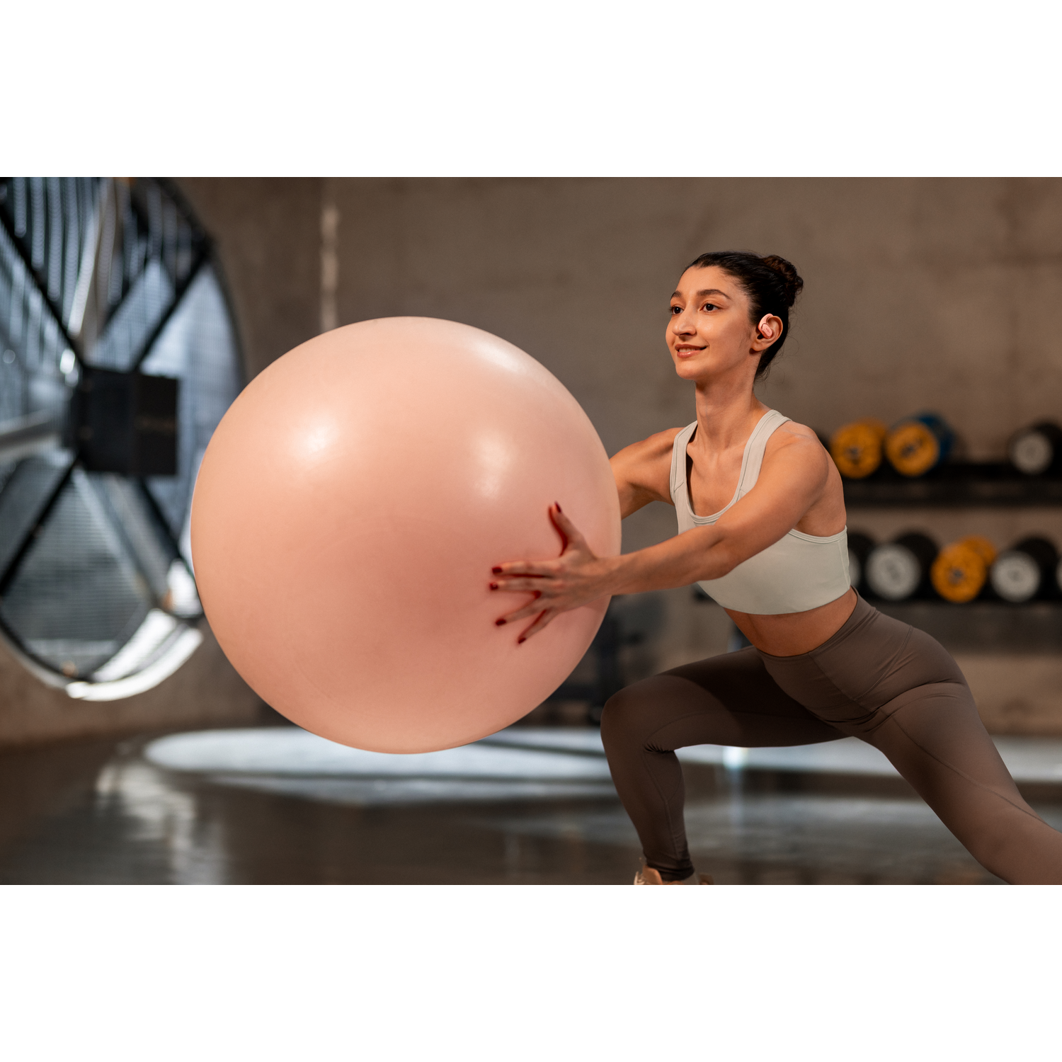 Woman in gym with pink exercise ball. She is wearing a white top and grey leggings. Earbuds are in.