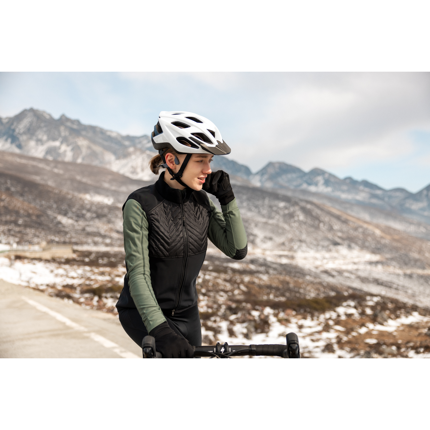 Woman in cycling gear with a helmet, earbuds, and a bike. Snowy mountains and sky in the background.