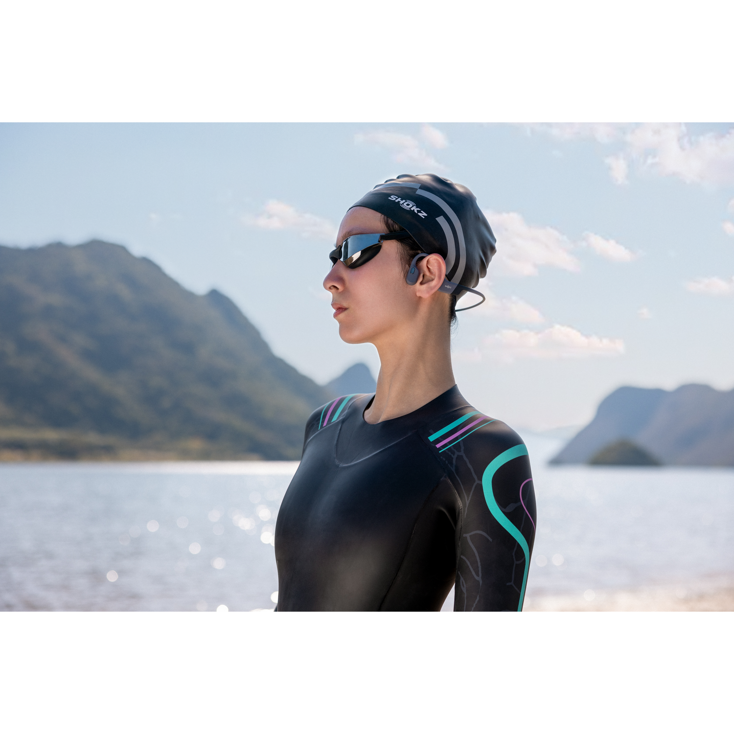 Woman in wetsuit and swim cap with goggles, near water. Mountains and sky in background.
