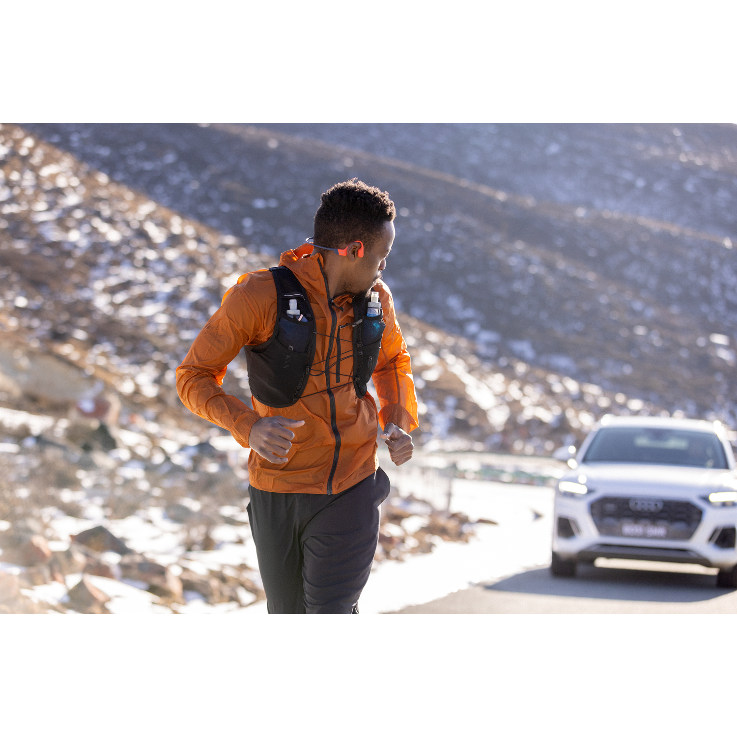 A runner in orange jacket runs on a road, with a car in the background.