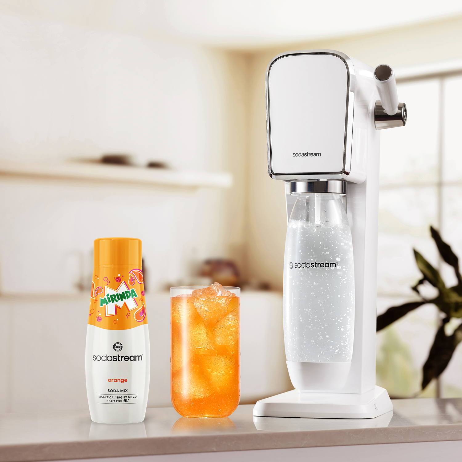 SodaStream setup: white machine, orange syrup, glass of orange drink with ice, white counter, and kitchen.