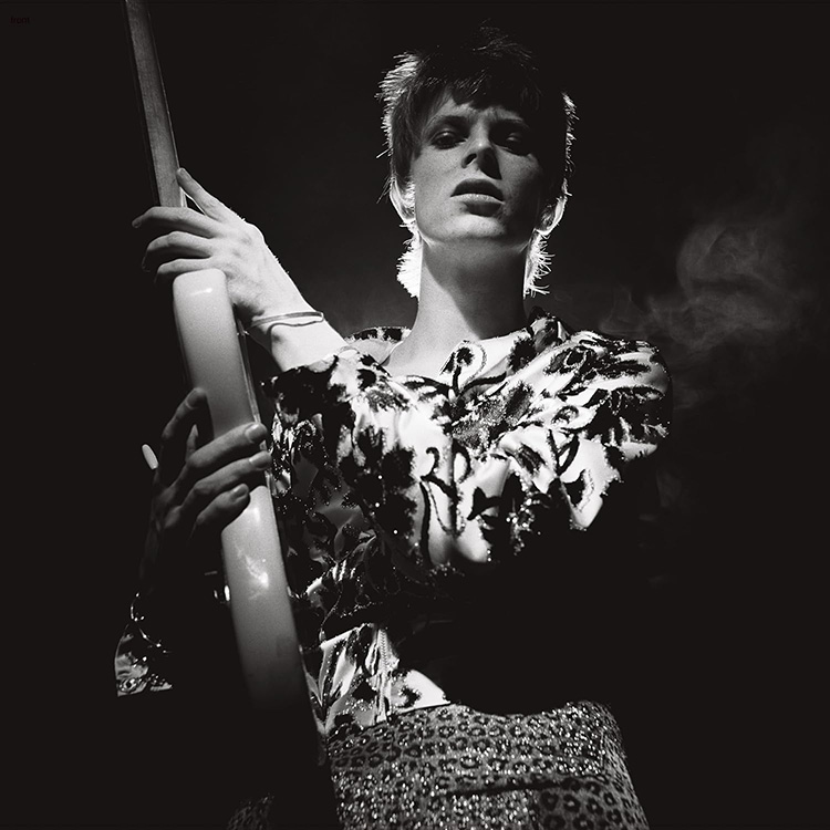 Bowie fekete-fehérben, fehér gitárt tartva, mintás felsőt és nadrágot visel.