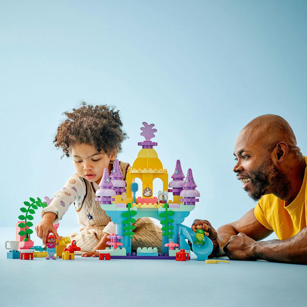Ein Kind und ein Mann spielen mit Lego-Steinen. Die Steine bilden eine Burgszene. Der Hintergrund ist blau.