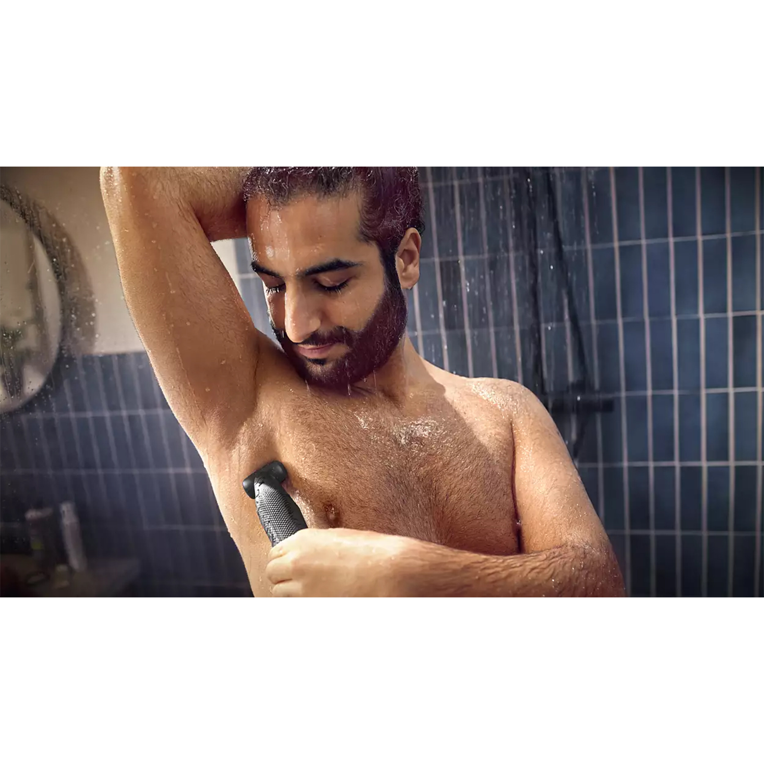 Uomo che si rade l'ascella con un rifinitore sotto la doccia. Acqua e piastrelle blu sullo sfondo.