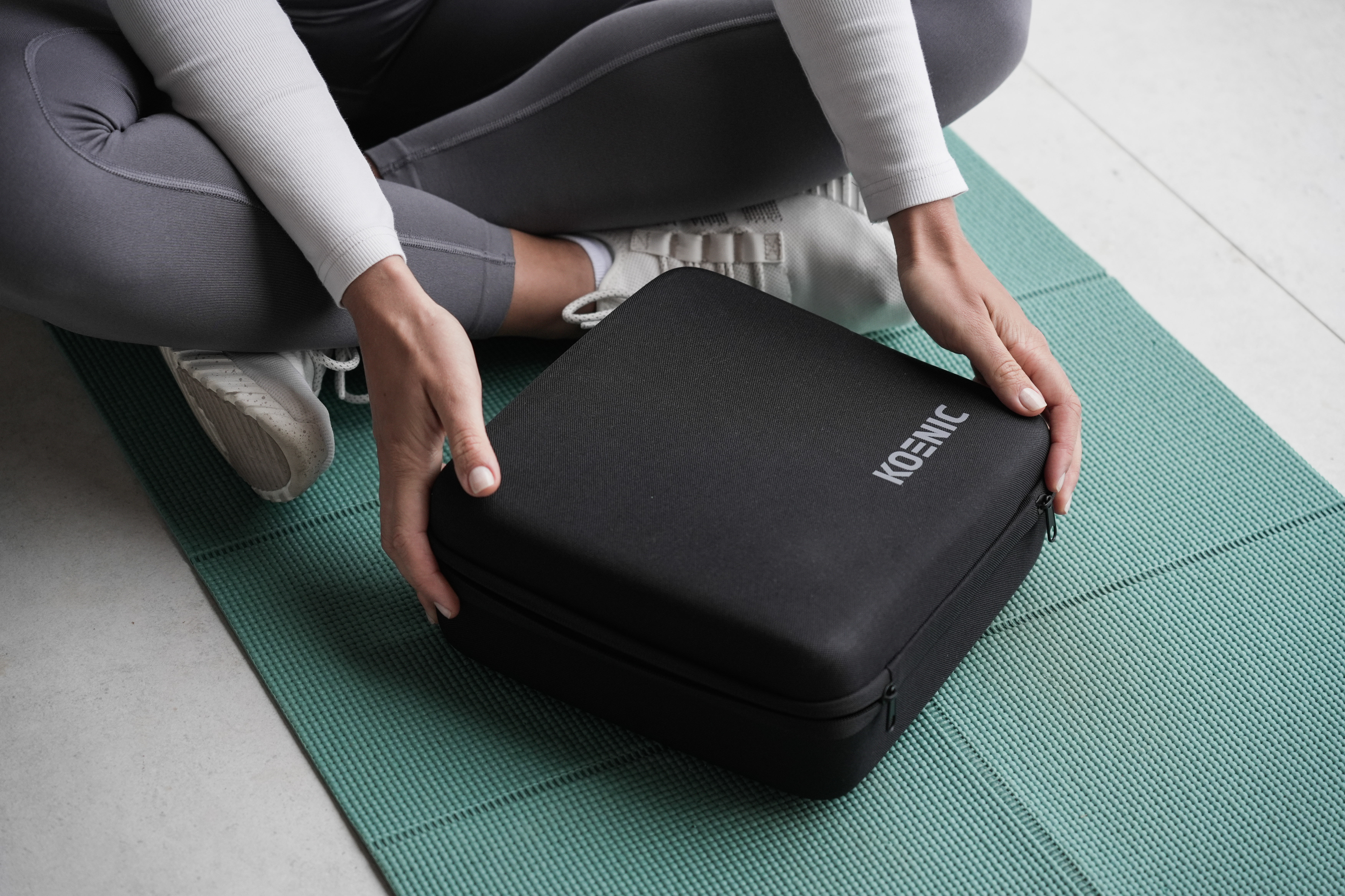 Person hält eine schwarze Koenic-Tasche auf einer grünen Yogamatte. Weiße Turnschuhe sichtbar. Innenraum.