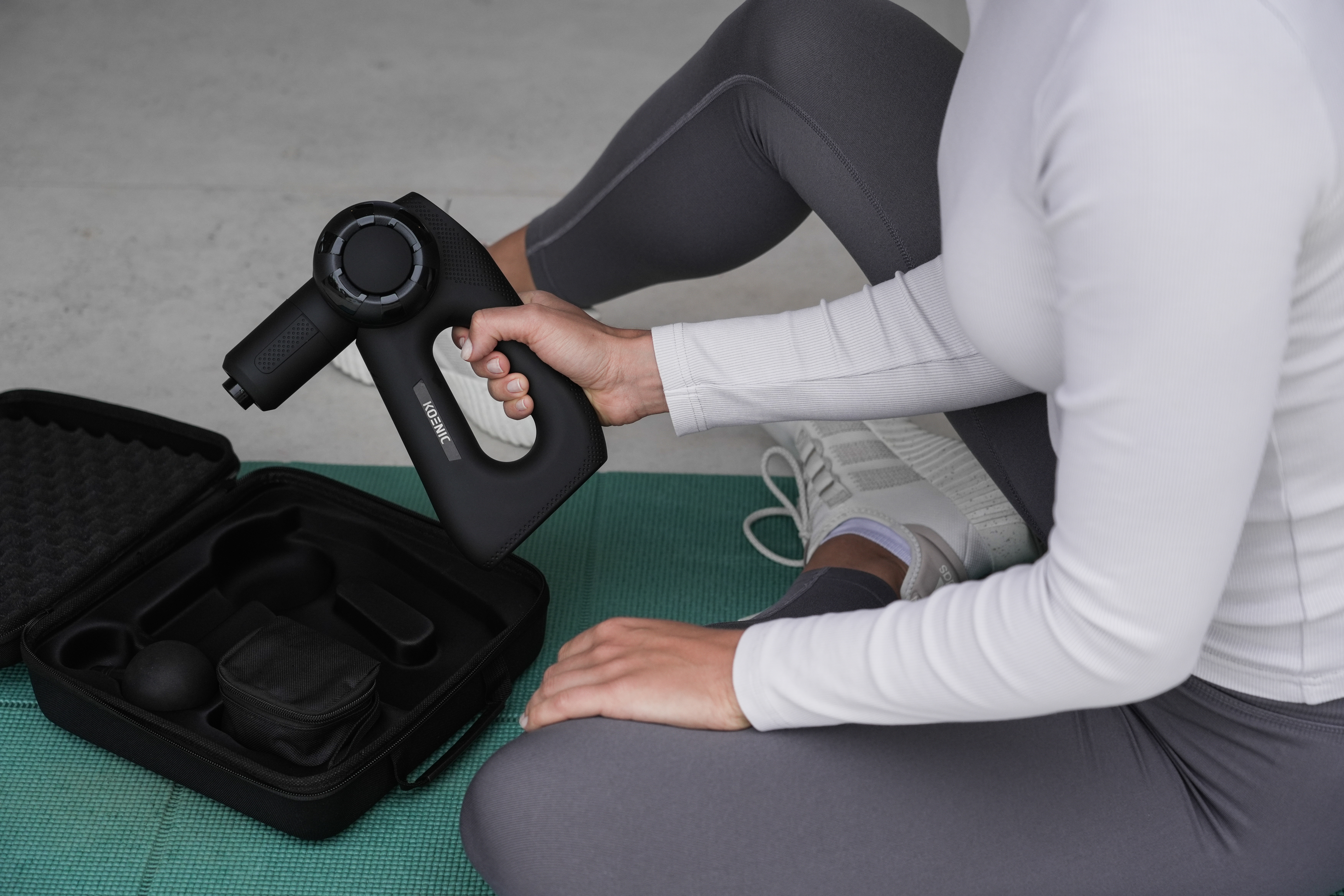 Eine Person hält eine schwarze Massagepistole, mit Zubehör und einem Koffer im Hintergrund.
