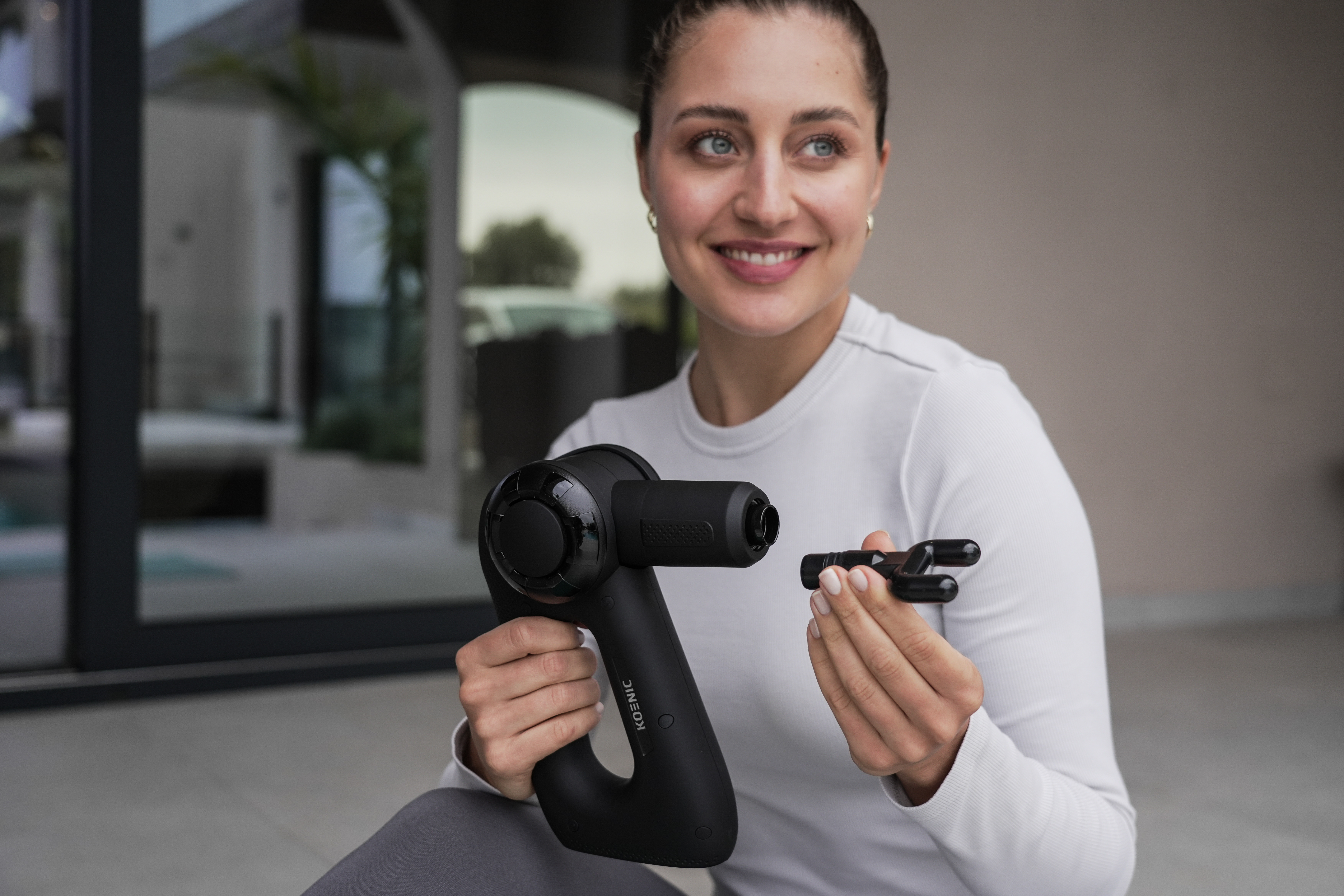 Eine Frau hält eine schwarze Massagepistole und einen schwarzen Aufsatz und lächelt in die Kamera.