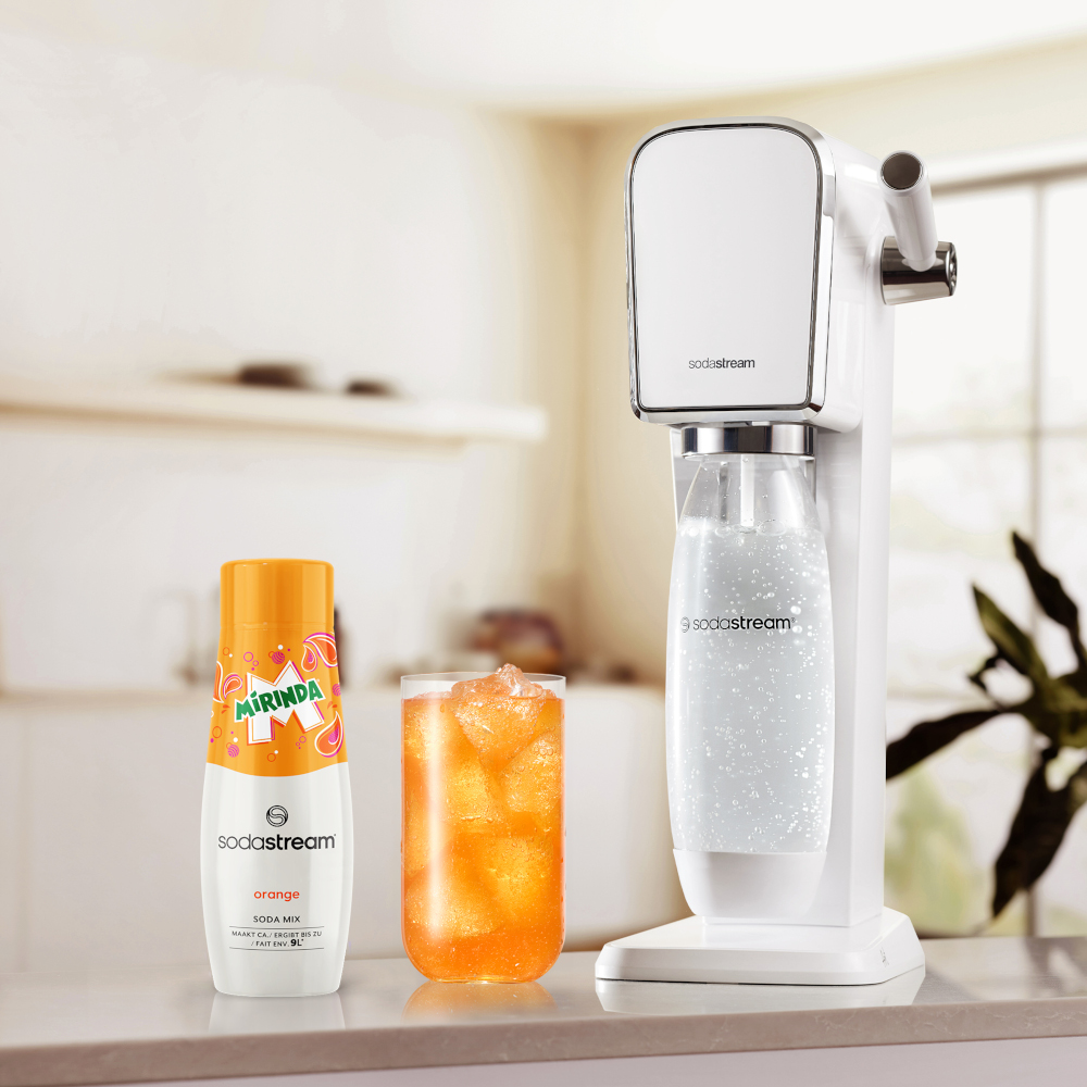 A white SodaStream machine, a bottle of orange flavor and a glass of orange soda with ice.