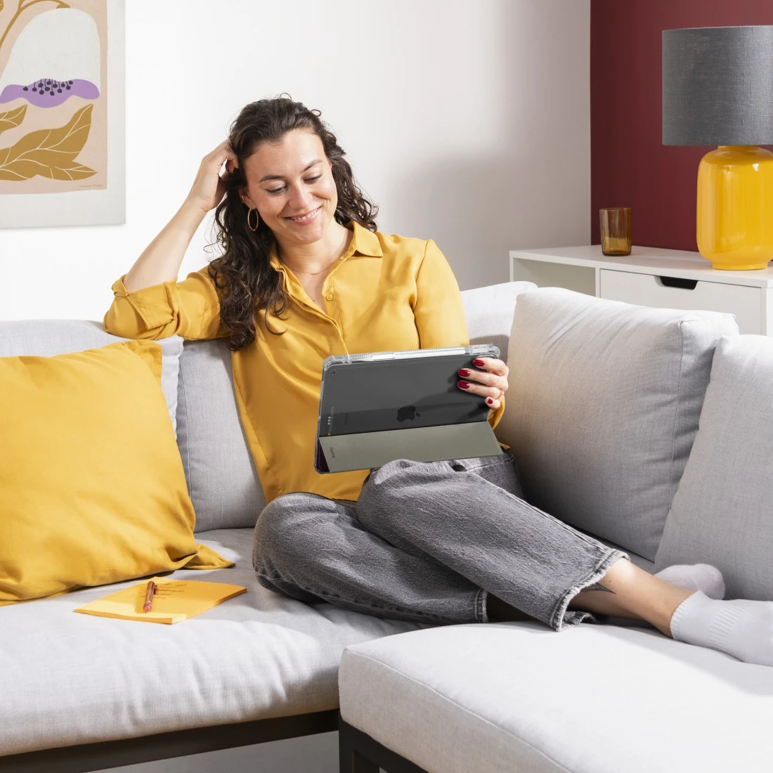 Donna su un divano che usa un iPad. Indossa una camicia gialla e pantaloni grigi. C'è un quaderno accanto.