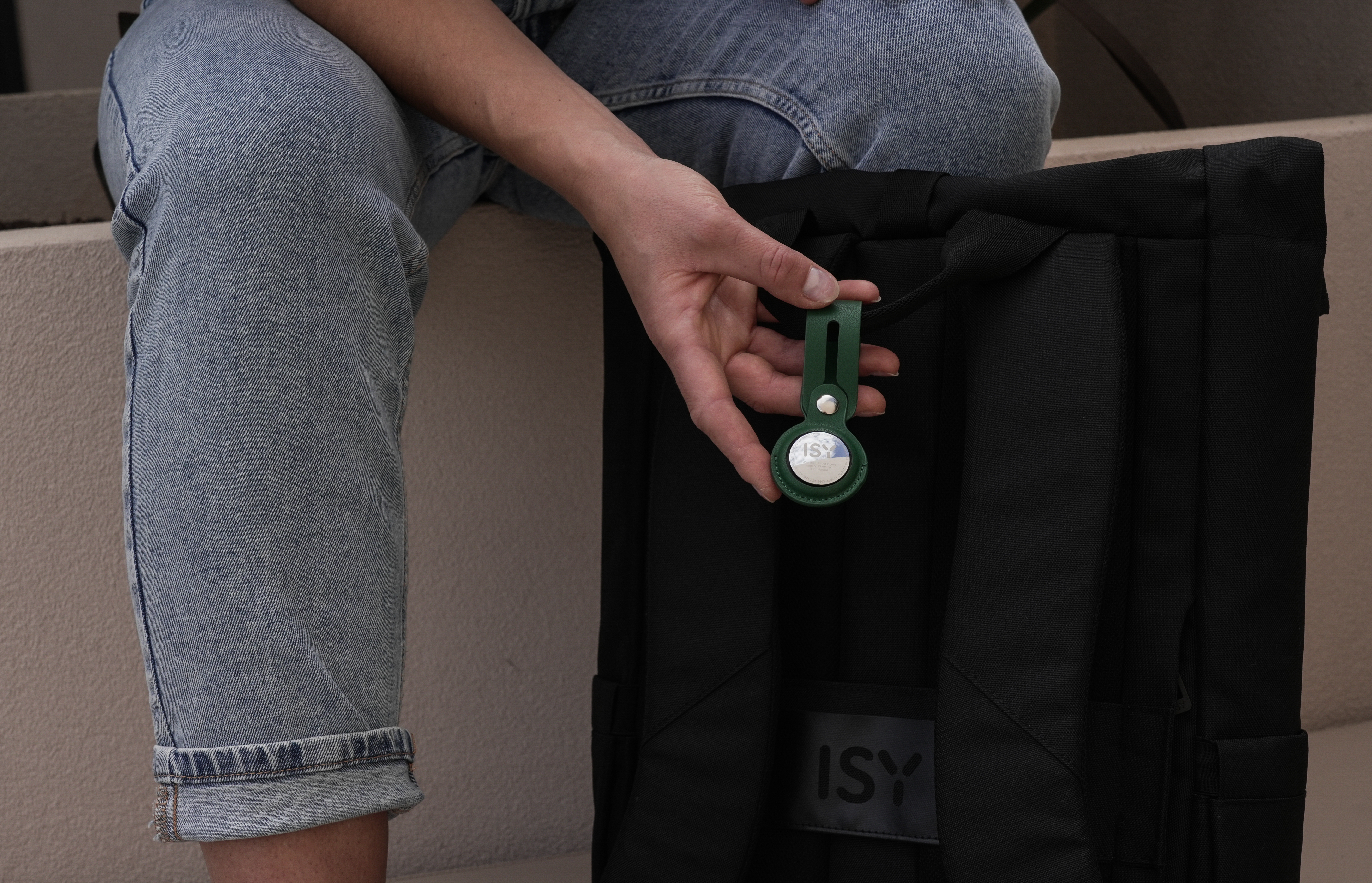 Die Hand einer Person hält ein grünes AirTag, das an einem schwarzen Rucksack befestigt ist. Jeans sind auch sichtbar.