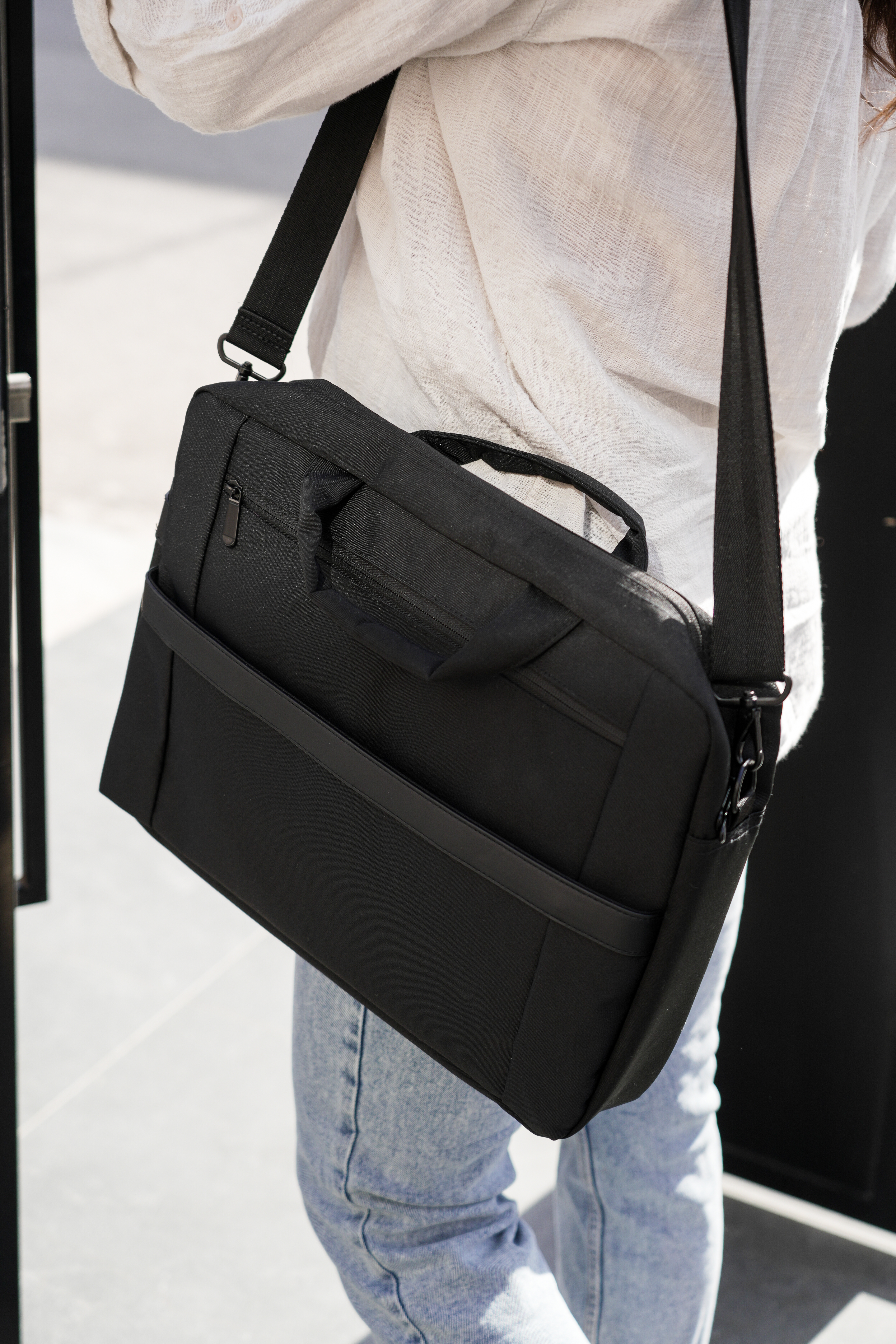 Person trägt eine schwarze Laptoptasche. Sie tragen ein weißes Hemd und Jeans, stehen draußen.