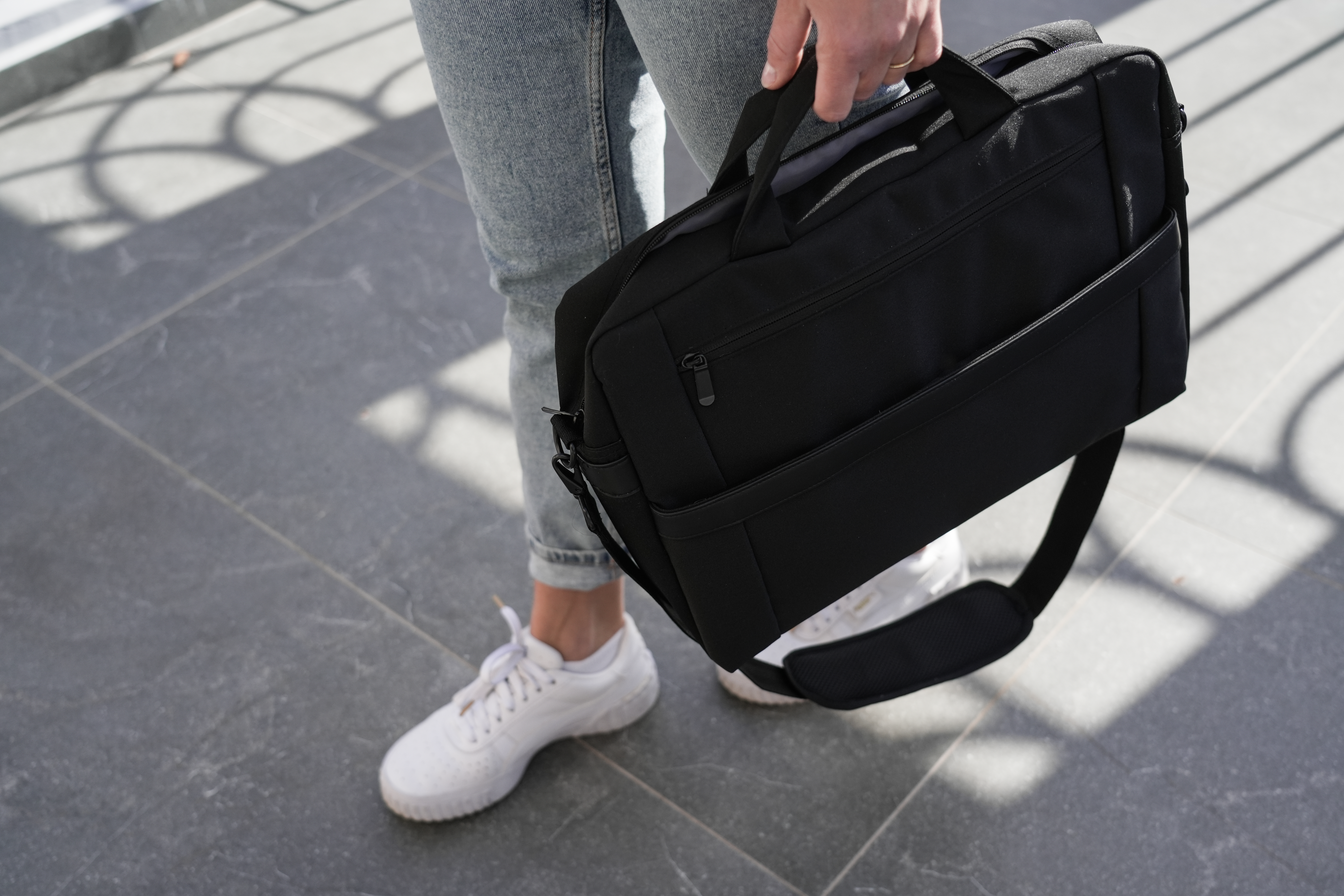 Eine Person in Jeans und weißen Turnschuhen hält eine schwarze Tasche mit Schultergurt. Die Tasche ist rechteckig.
