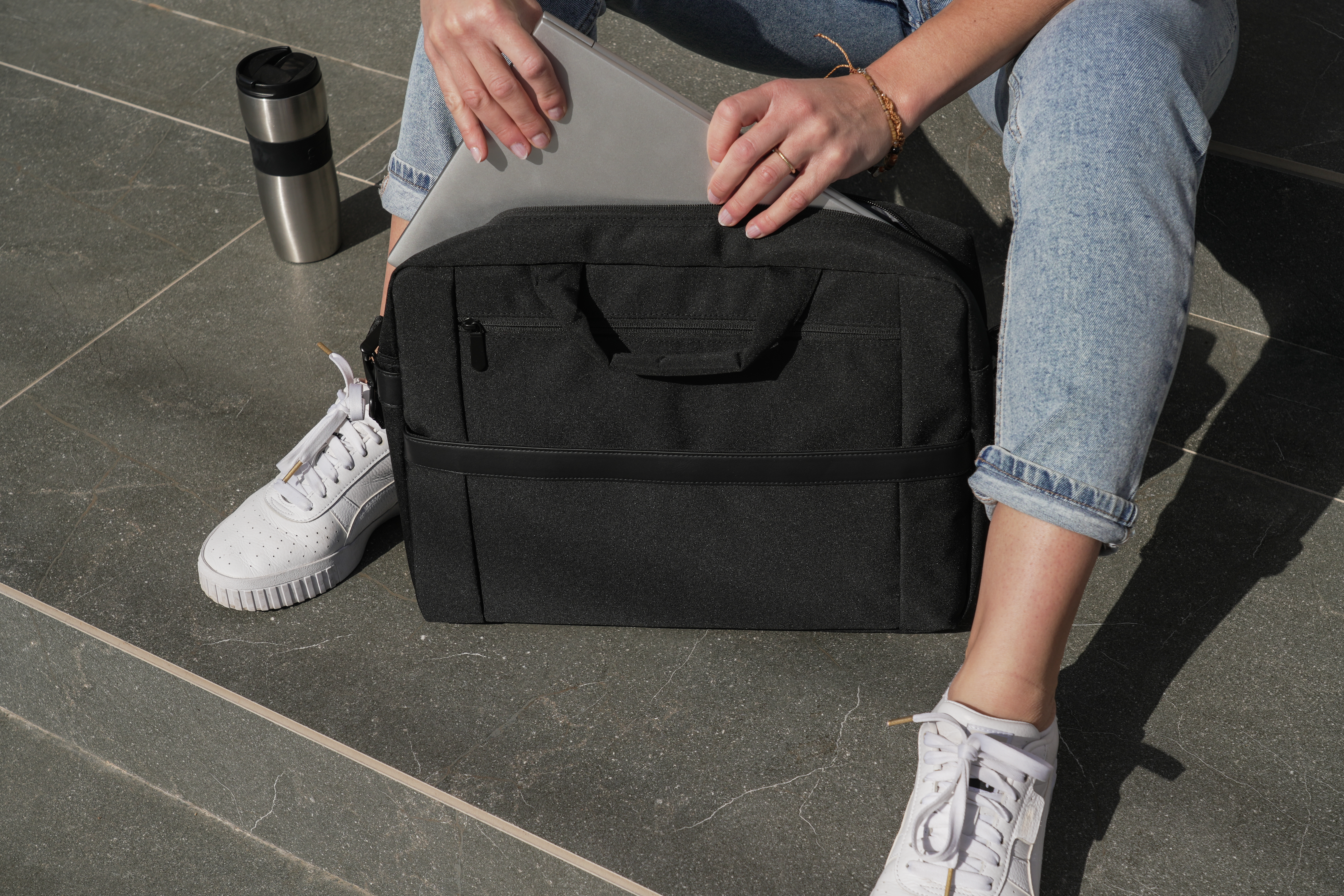 Person öffnet eine schwarze Tasche, neben einer silbernen Thermoskanne und weißen Turnschuhen. Sie trägt Jeans und hat einen Ring.