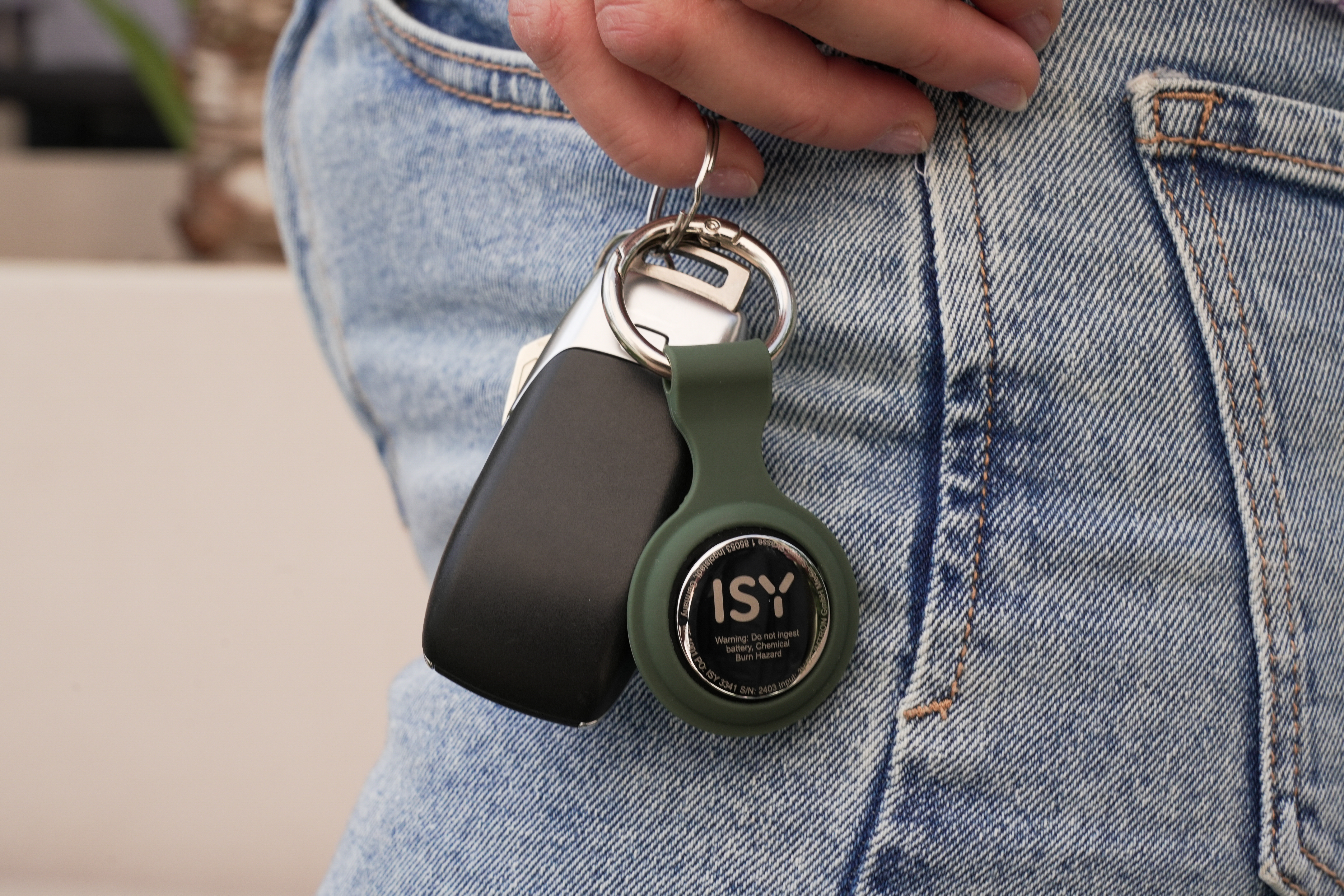 Hand hält Schlüssel und einen ISY-Tracker an einer Jeans-Tasche fest. Schlüssel sind schwarz und silber. Tracker ist olivgrün.
