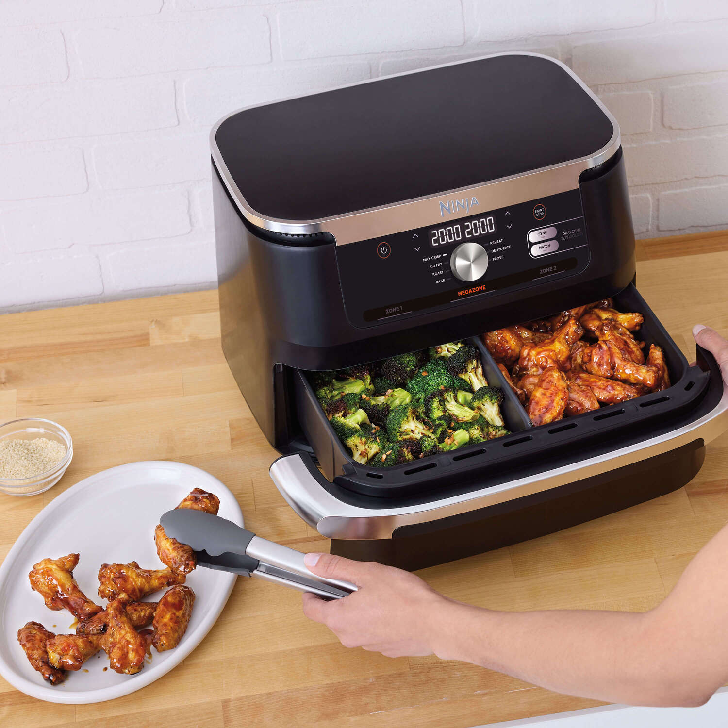 Zwarte Ninja airfryer met bakken met eten, een bord en een tang op een houten oppervlak.