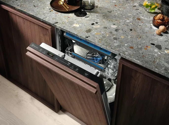 Zmywarka z otwartymi drewnianymi drzwiami, odsłaniająca stojaki z naczyniami. Granitowy blat w tle.