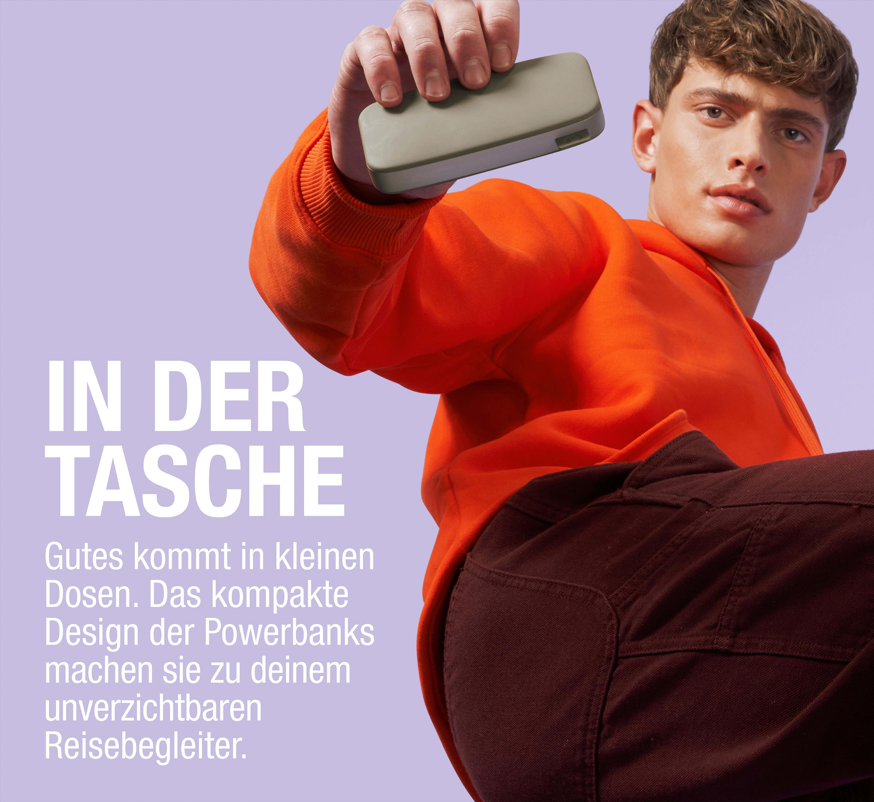 Ein Mann hält eine Powerbank, trägt einen orangefarbenen Hoodie und braune Hose.