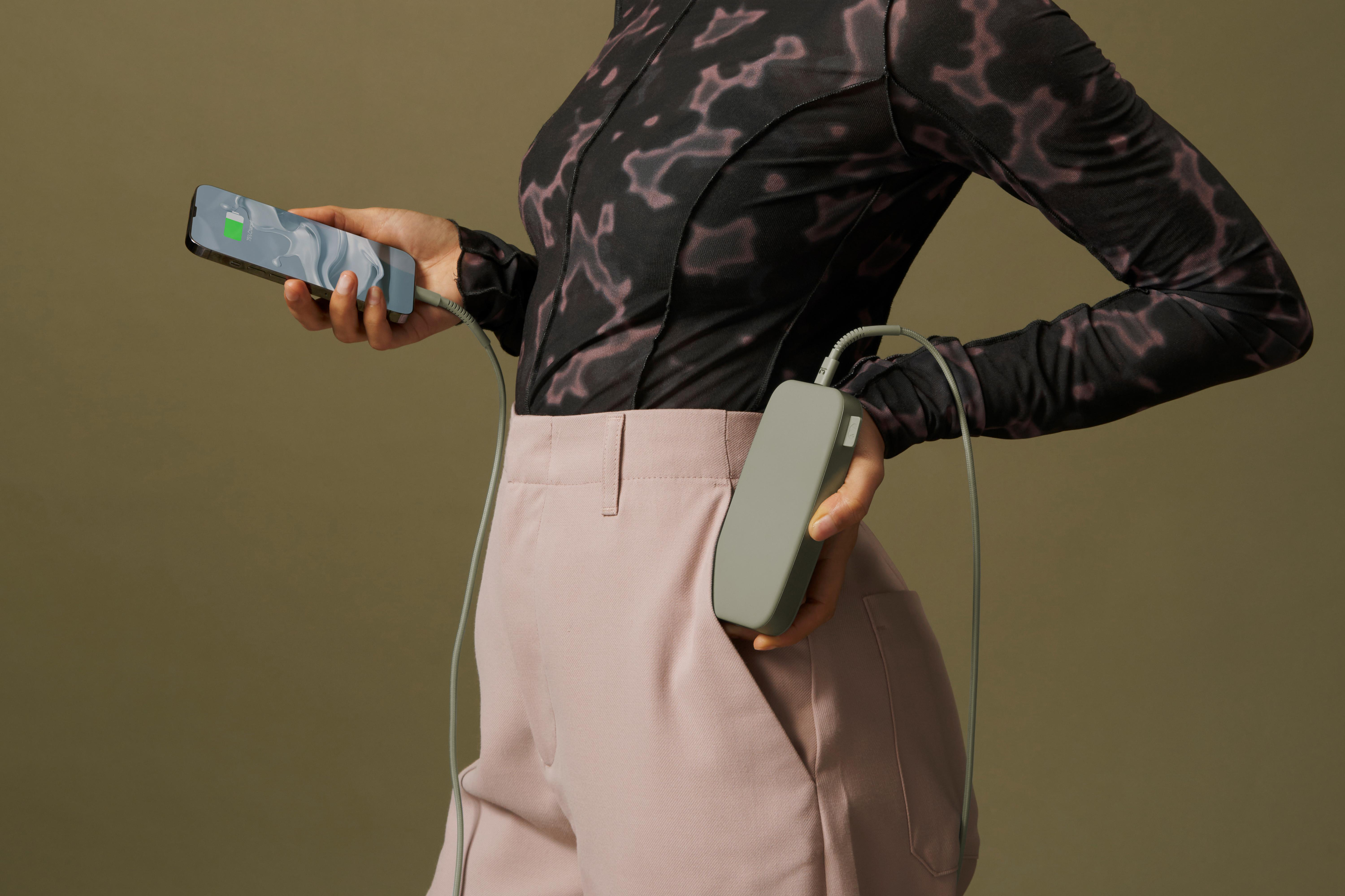 Person in pinker Hose hält ein Telefon, das von einer Powerbank in ihrer Tasche geladen wird. Der Hintergrund ist olivgrün.