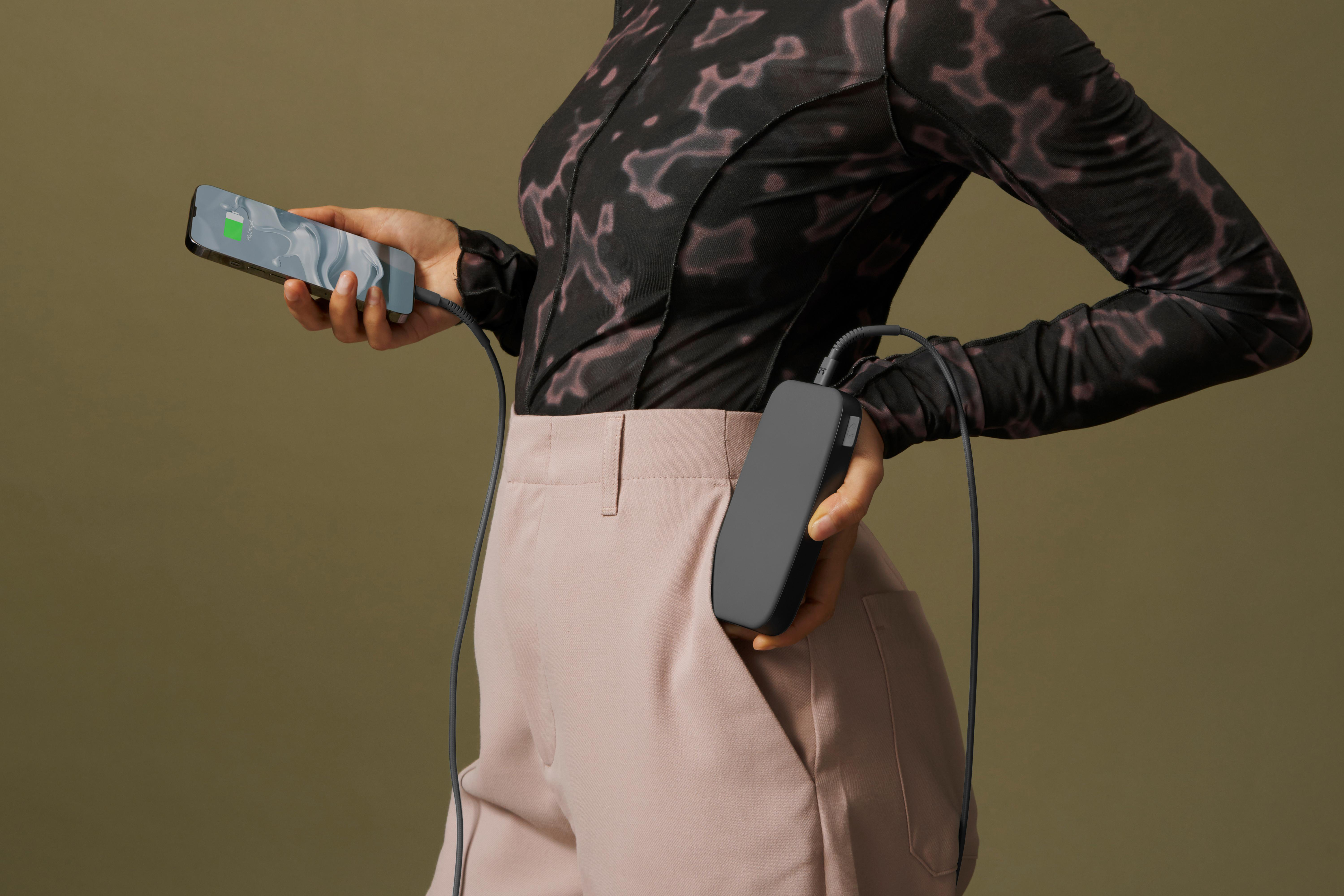 Person hält ein Telefon, das von einer Powerbank in ihrer Tasche geladen wird. Der Hintergrund ist eine neutrale grüne Farbe.