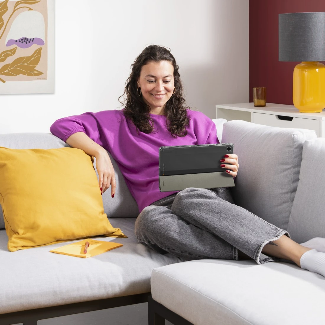 Frau auf Couch mit Tablet. Gelbes Kissen und Lampe neben ihr.