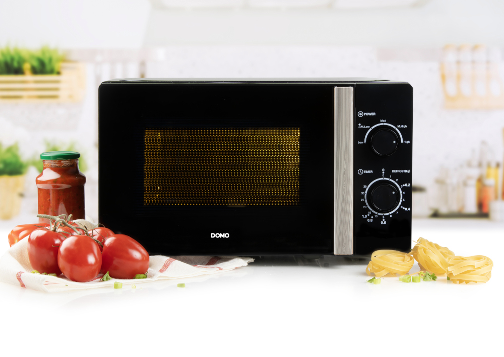 Un micro-ondes noir avec des tomates et des pâtes au premier plan. Il y a aussi un pot de sauce.