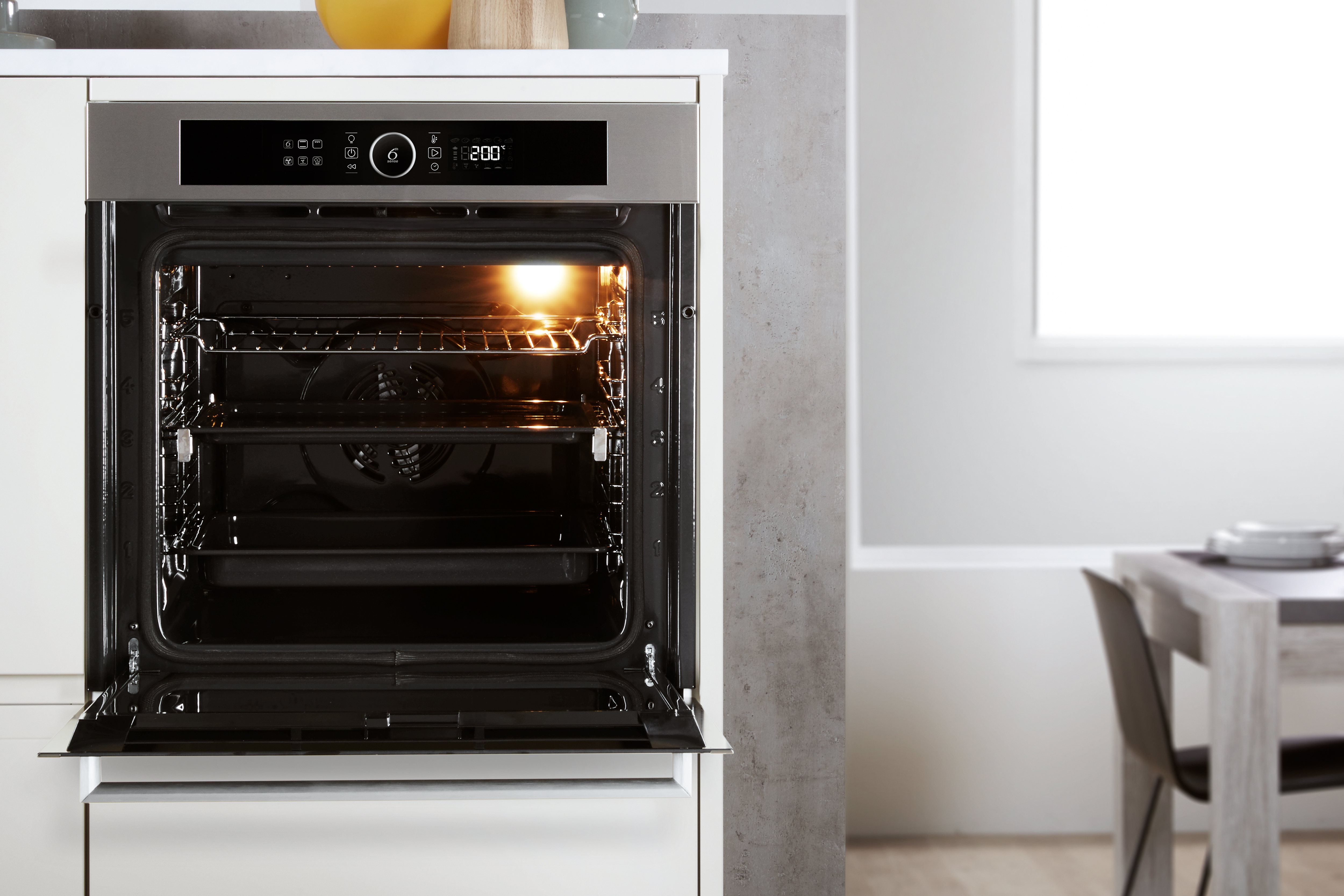 Horno abierto con luz interior brillante. Armarios blancos, mesa y ventana en el fondo.