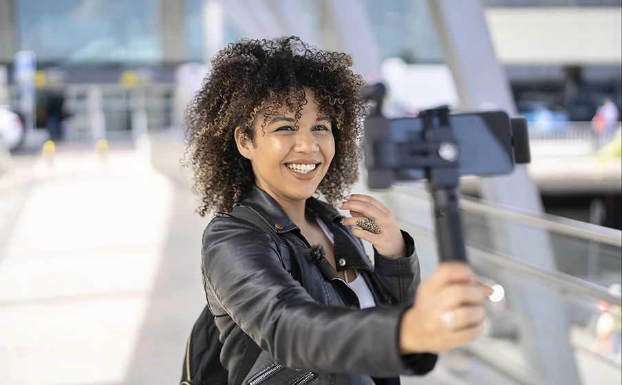 Kıvırcık saçlı bir kadın, selfie çubuğunda bir telefon tutarken gülümsüyor.