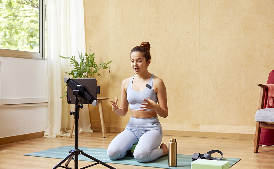 Bir kadın, bir yoga matının üzerinde diz çöküyor, bir tripod üzerindeki bir cihazla konuşuyor. Bir yoga stüdyosunda.