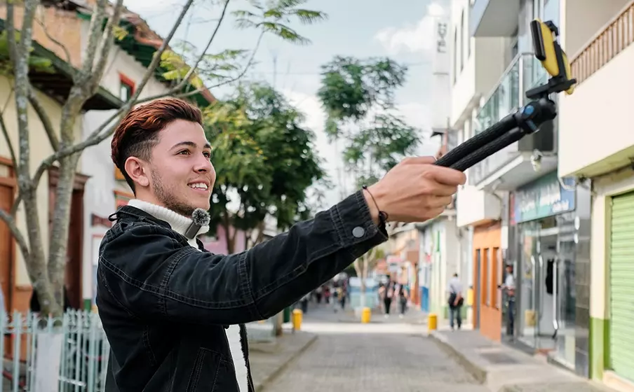 Adam, bir tripod üzerine takılı bir akıllı telefonla sokakta kendini filme alıyor, gülümsüyor.