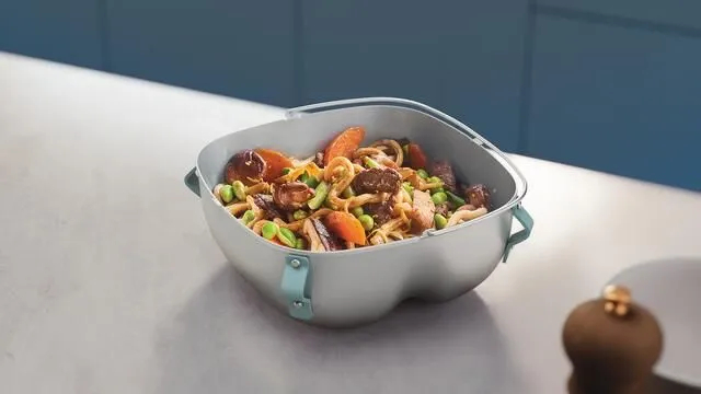 Pojemnik z makaronem, warzywami i mięsem. Stoi na jasnoszarym blacie kuchennym.