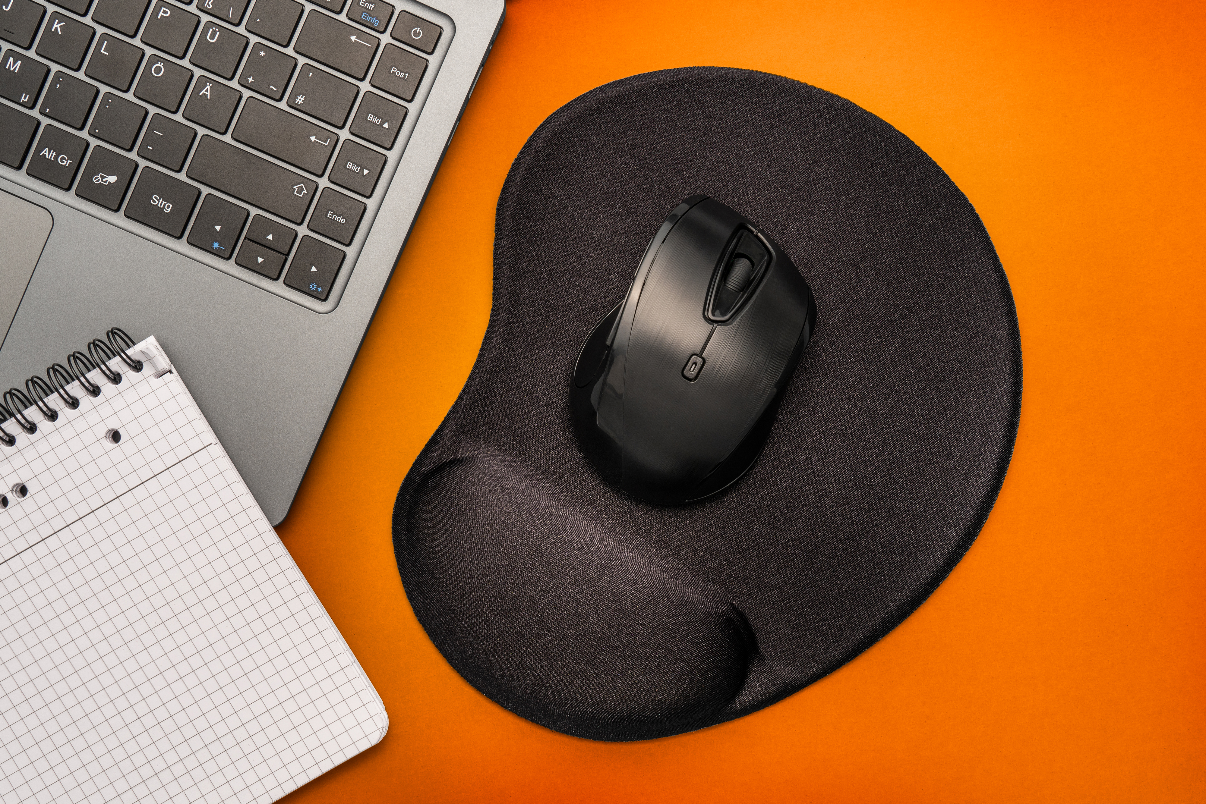 Espace de travail avec ordinateur portable, bloc-notes et souris ergonomique noire sur surface orange.