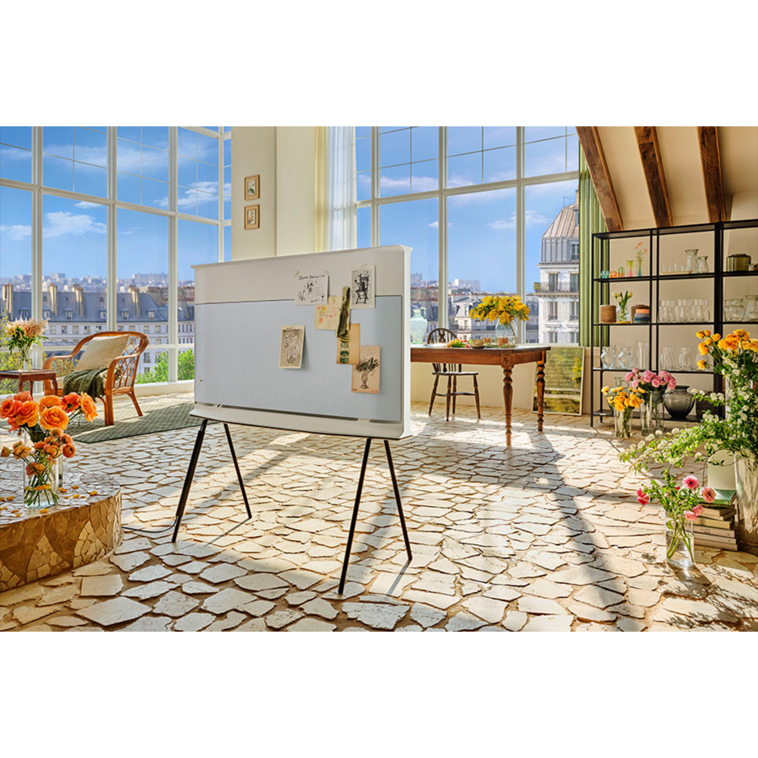 A modern living room with a TV on a stand. Large windows offer a city view. Flowers in vases and a unique floor.