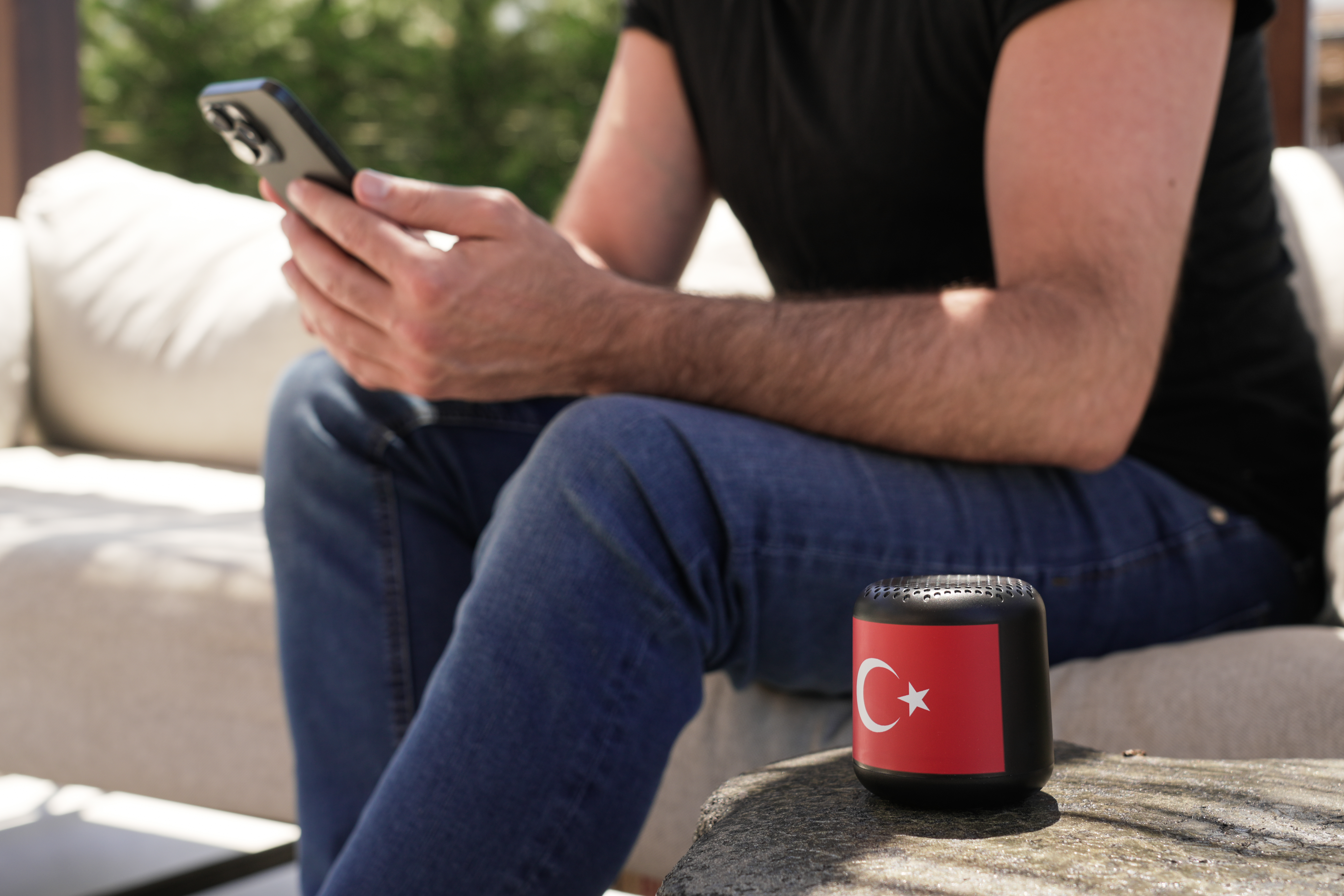 Ein Mann sitzt mit einem Telefon und einem kleinen Lautsprecher mit türkischer Flagge. Er ist draußen auf einem Sofa.