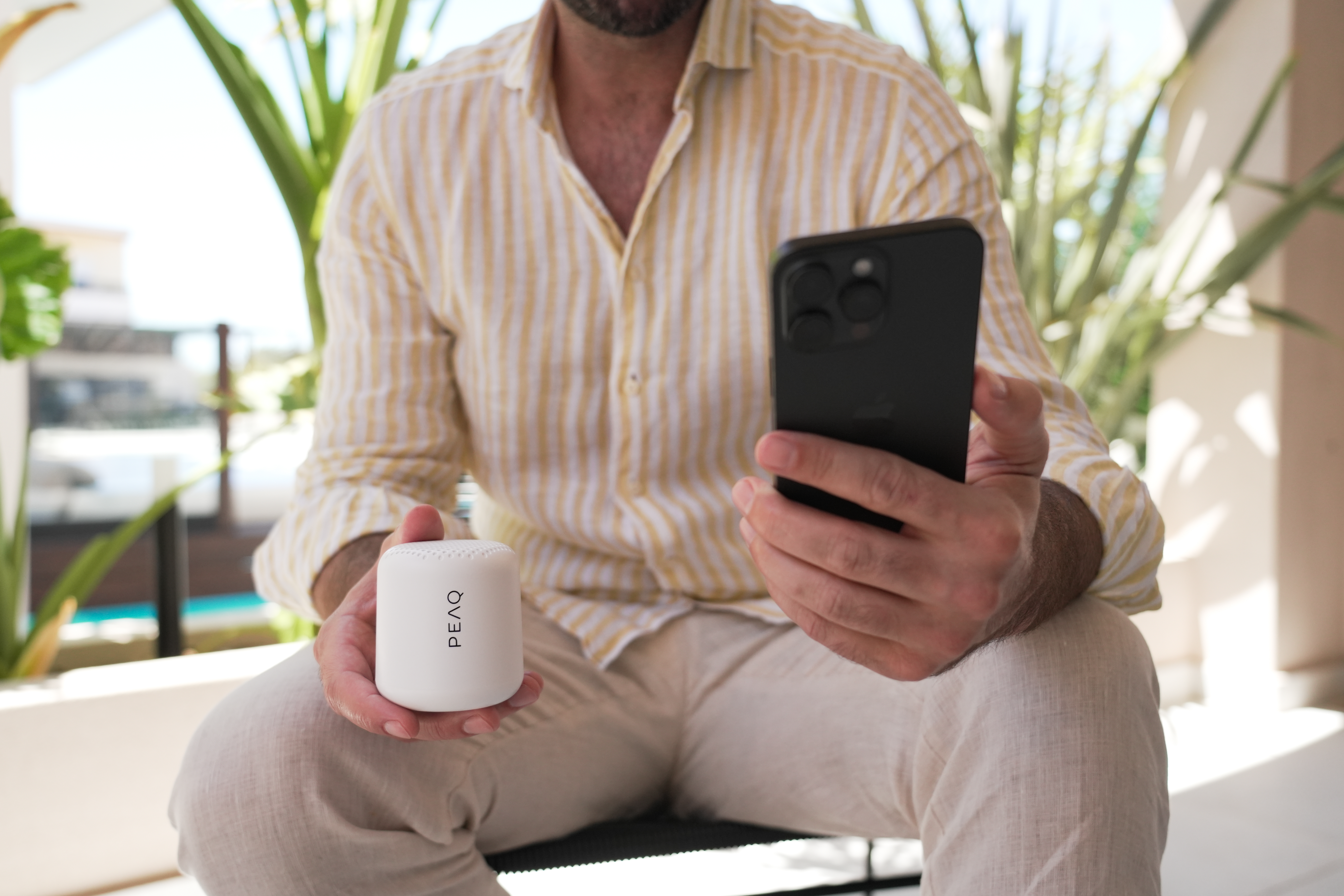 Ein Mann hält einen weißen Lautsprecher und ein schwarzes Telefon, sitzt draußen mit Grün im Hintergrund.